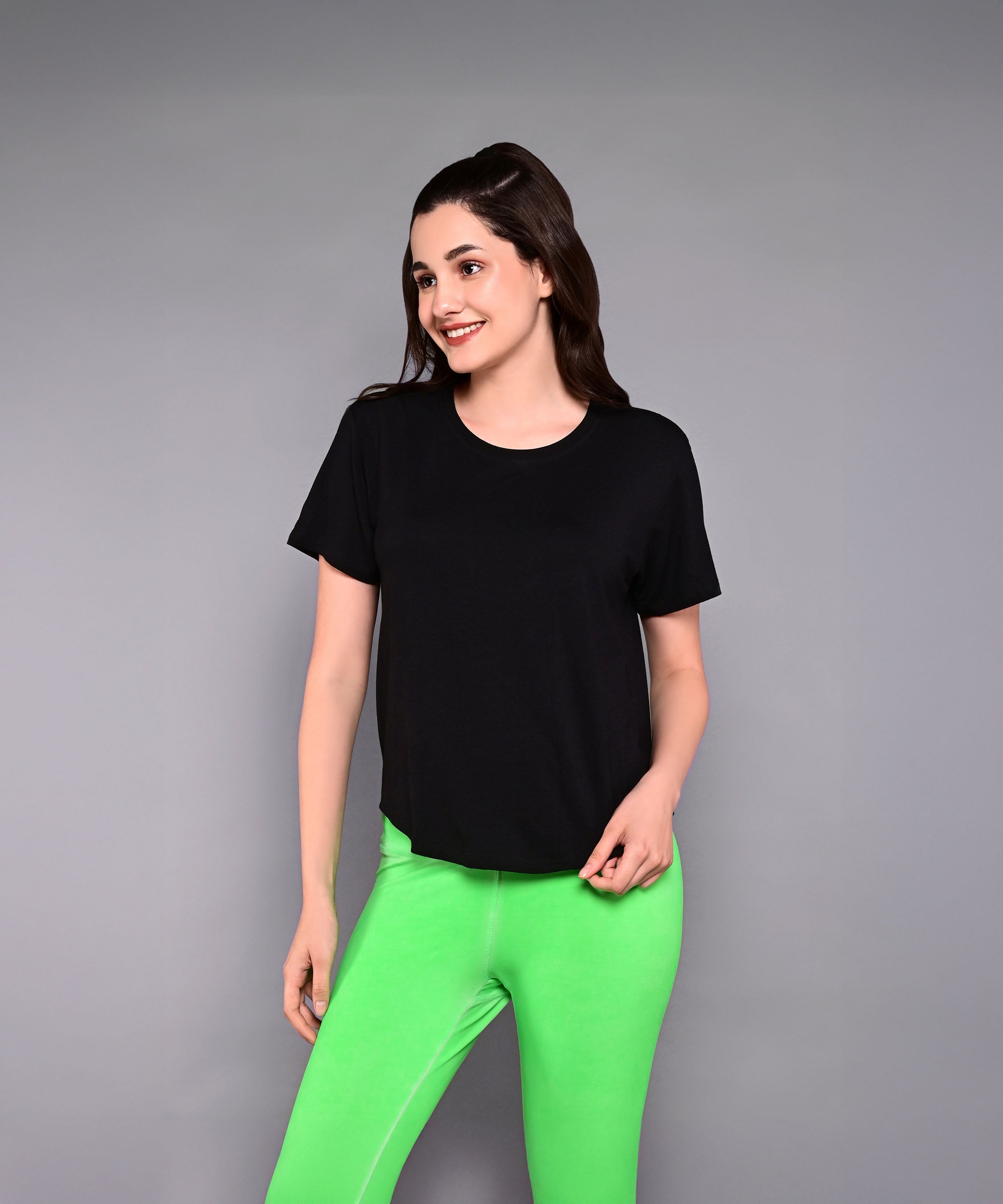 A woman stands against a plain gray background, smiling in the 'Pilates Has Your Back' Printed Tee and bright green pants.