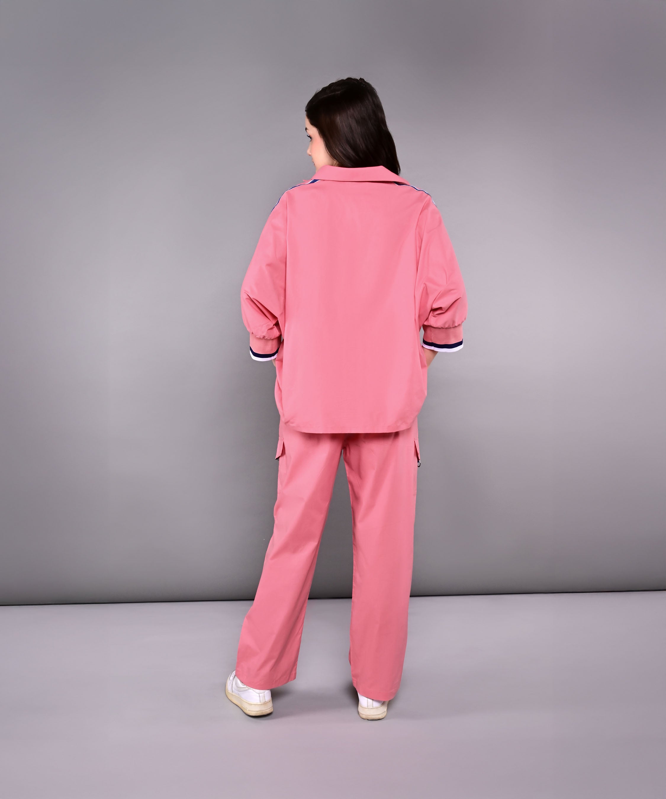 A woman stands with her back to the camera, wearing the Effortlessly Chic Set— a pink cotton co-ord featuring a batwing sleeve shirt and cargo track pant—against a gray background.
