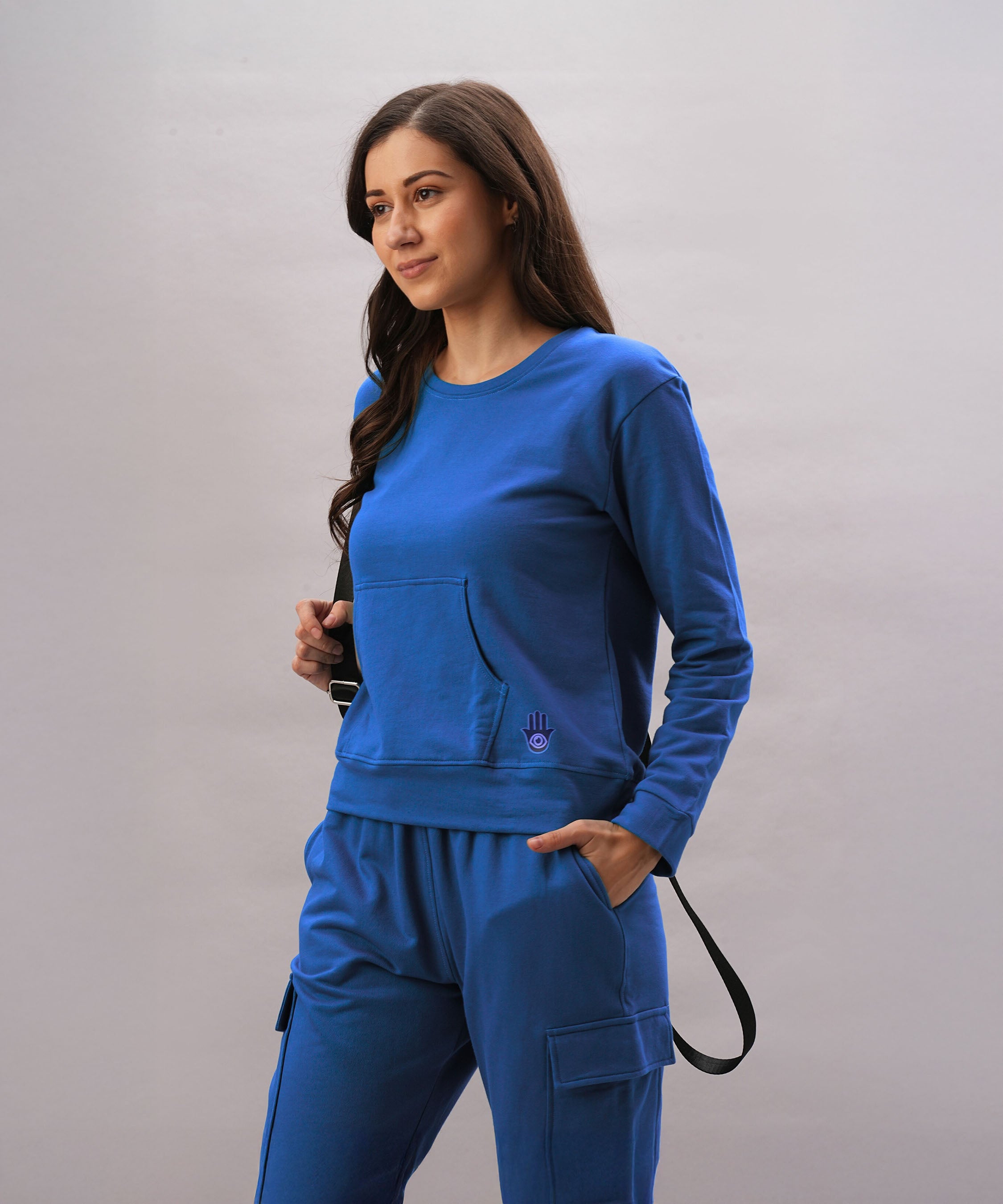A woman wears the Soft Royal Blue Trackset with cargo-style joggers, her hand in her pocket and holding a black strap, set against a light background.