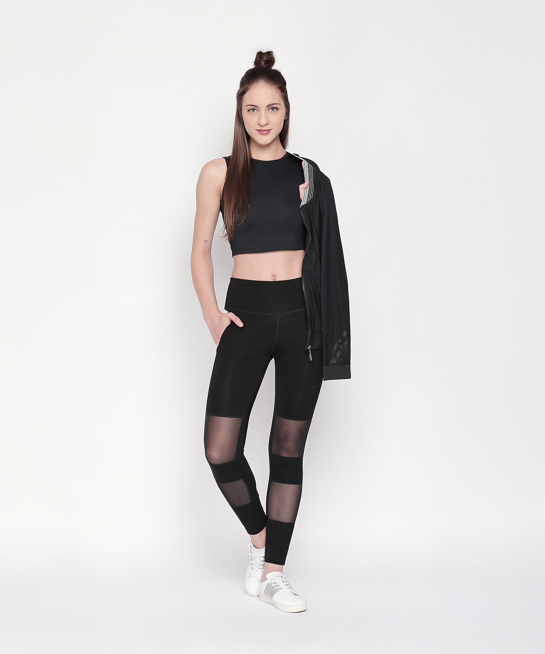 A woman wearing the Active Wear Crop Top in Black Colour, mesh-paneled leggings, and white sneakers, holding a black jacket and standing against a white background.