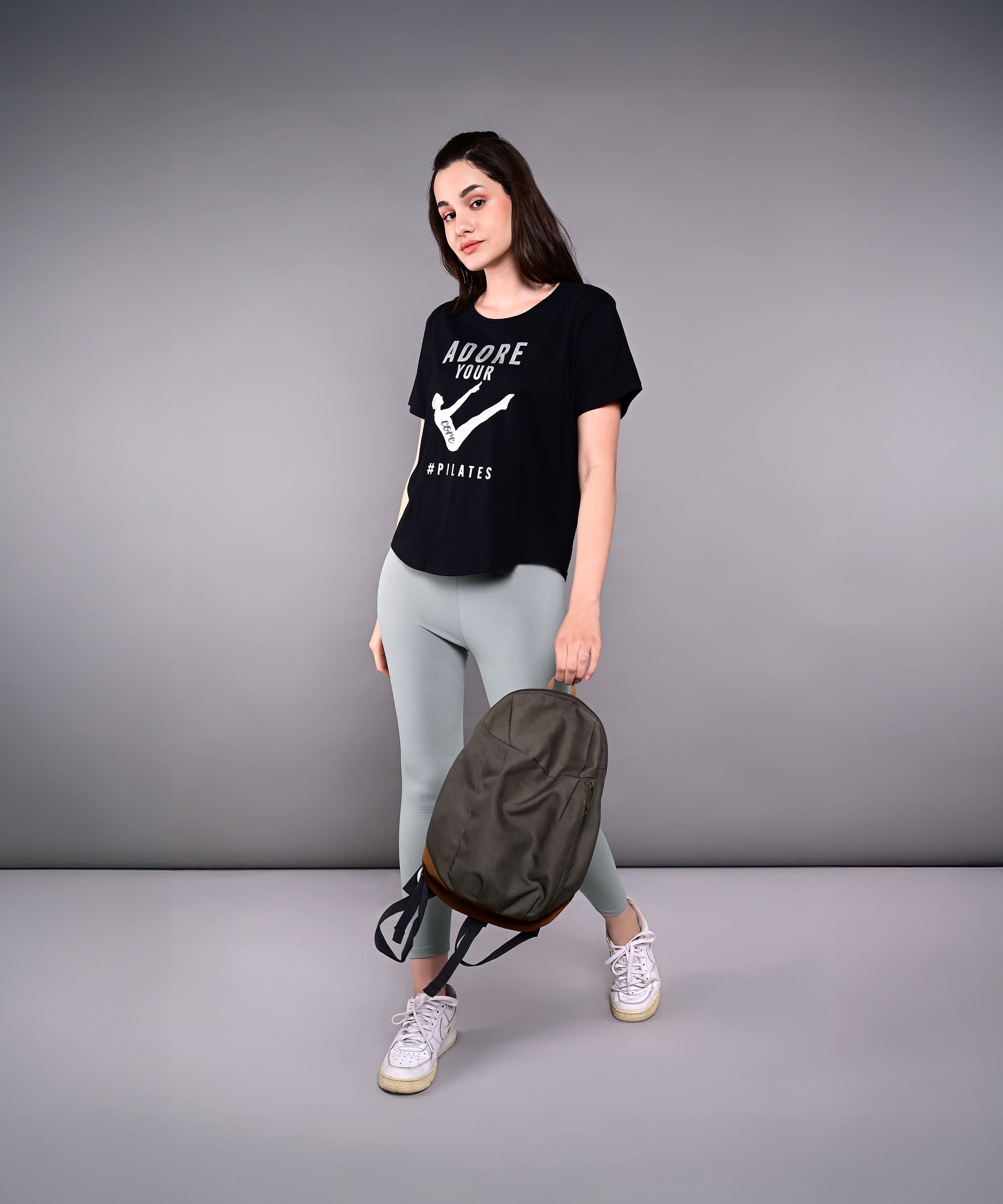 Woman in an 'Adore Your Core' printed tee and light leggings, holding a backpack, stands against a plain gray background.
