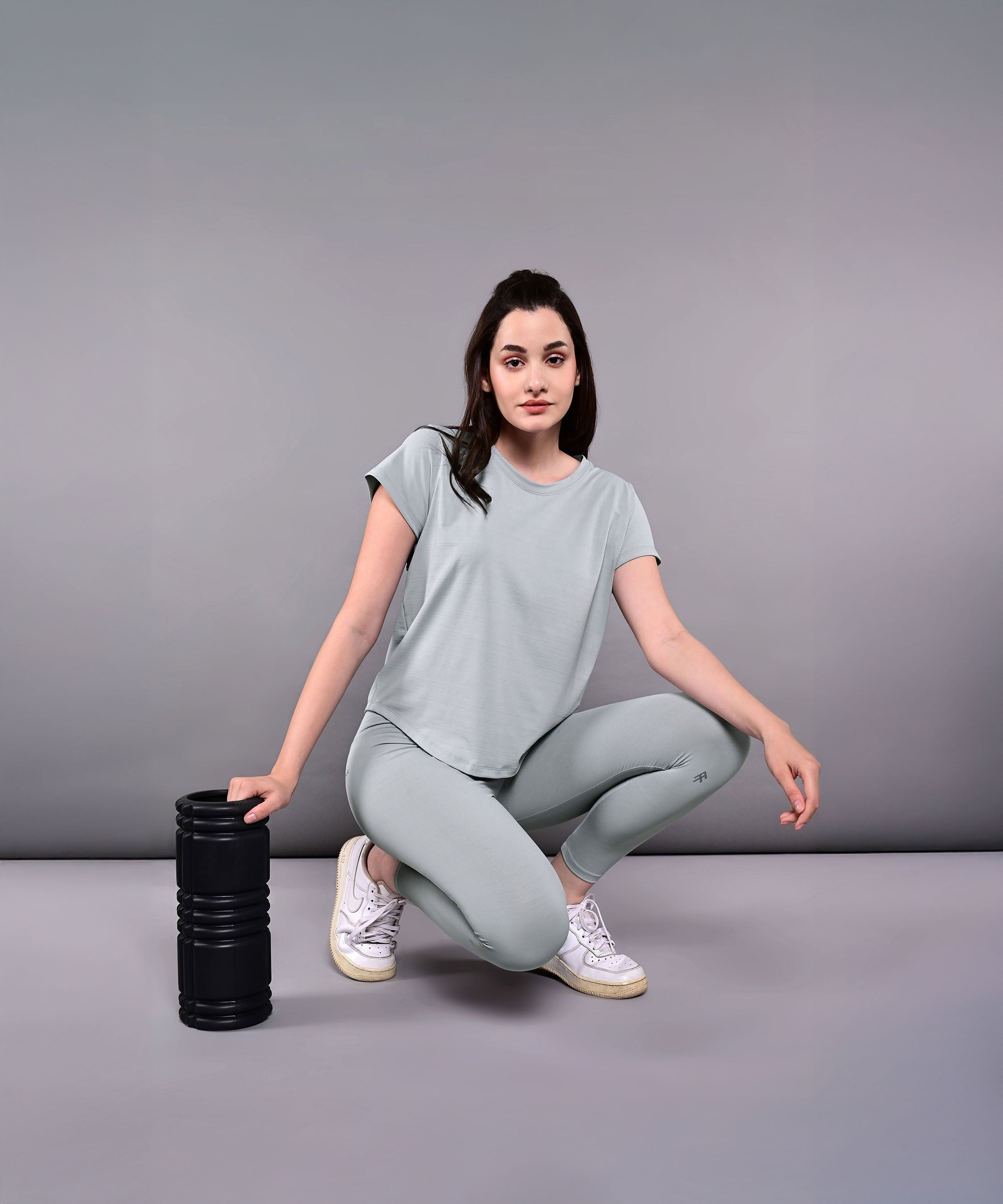 A woman wearing the Dynamic Grey Performance Set squats on the floor beside a black foam roller against a plain background.