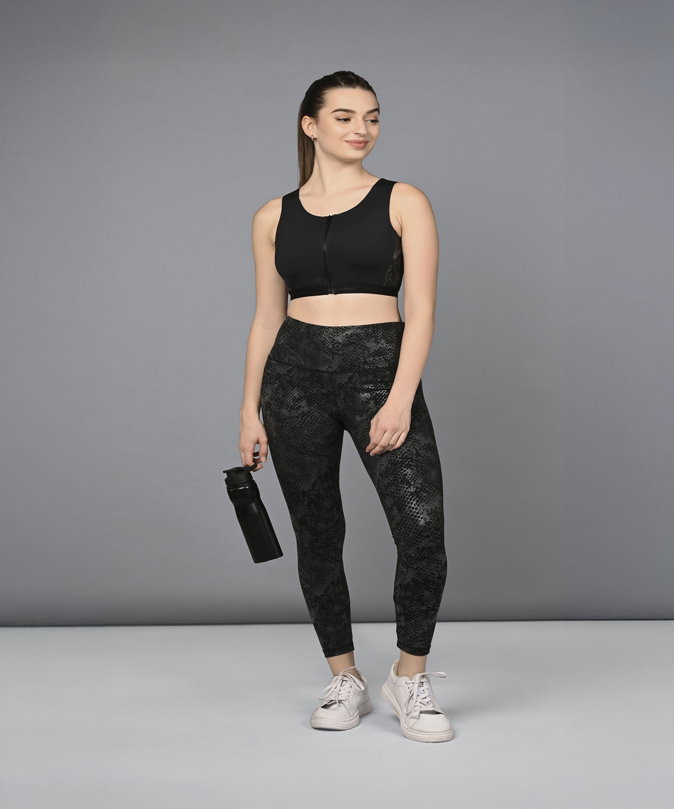 Woman wearing the Zipped Sports Bra & Metallic Legging activewear set, holding a water bottle and smiling while standing against a plain gray background.