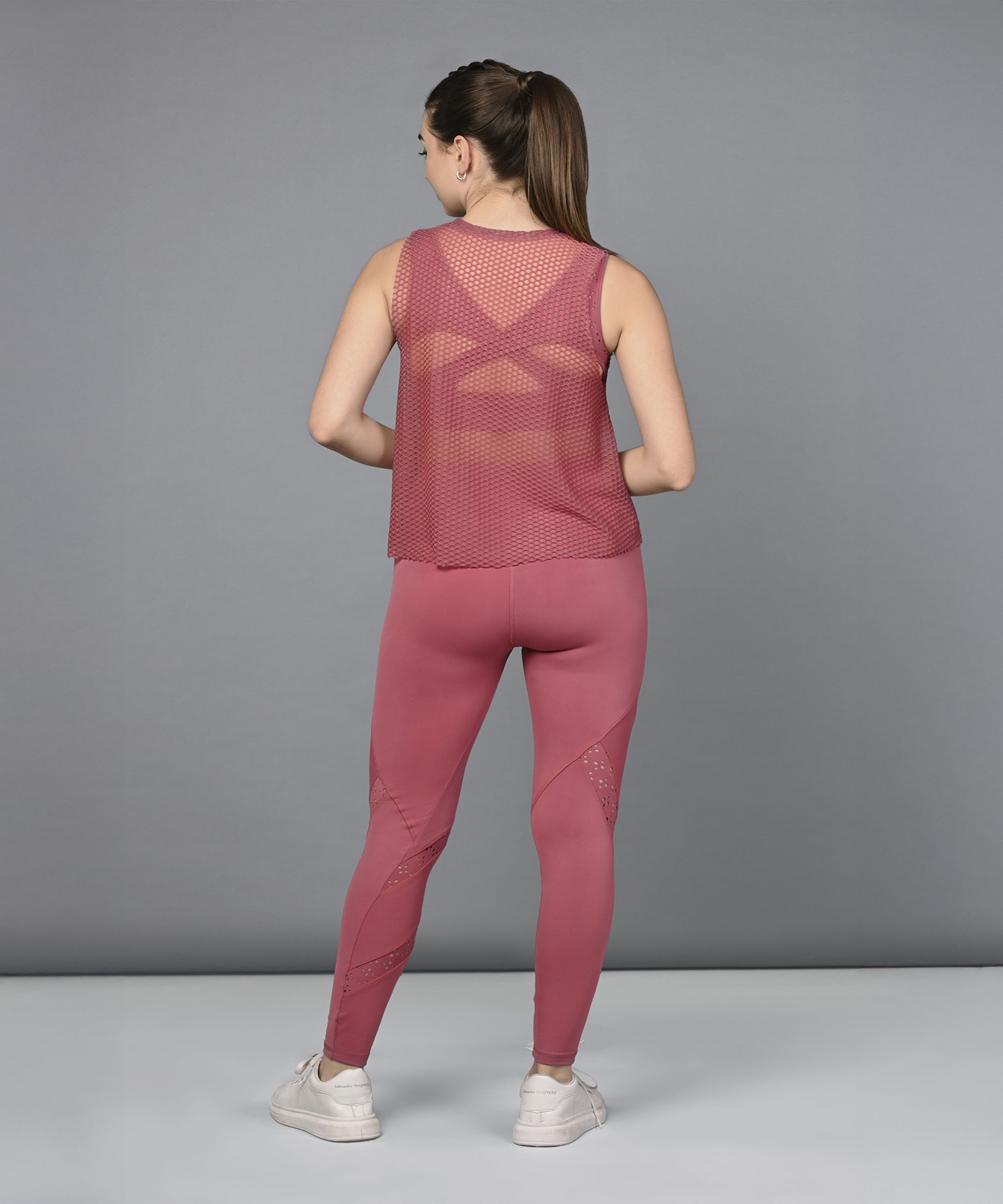 A woman stands facing away in the Peony Pink Netted Top, displaying its mesh design and honeycomb self pattern against a gray background.