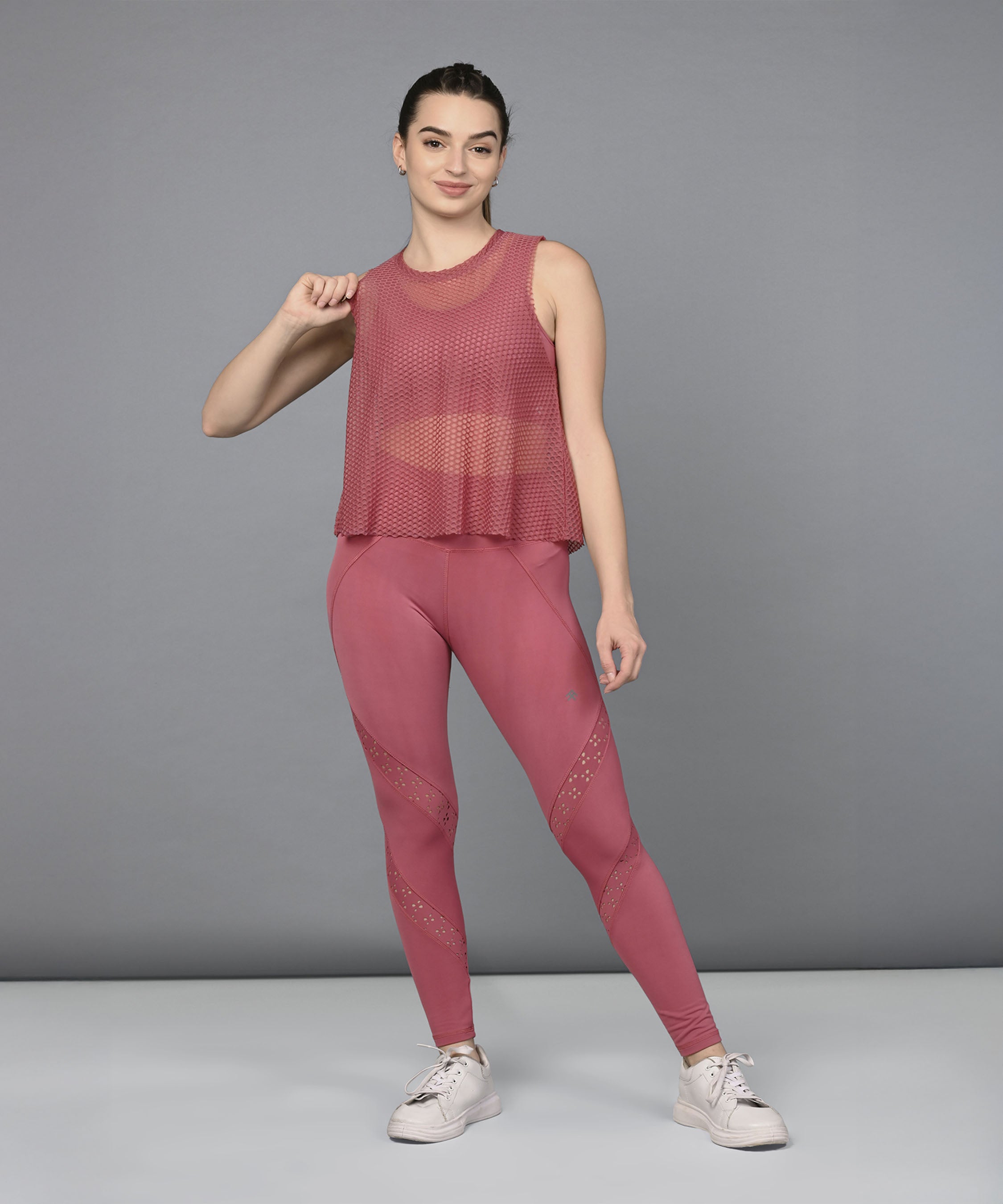 A woman poses confidently against a gray background, showcasing the Peony Pink Netted Top paired with white sneakers for a chic, sporty look.