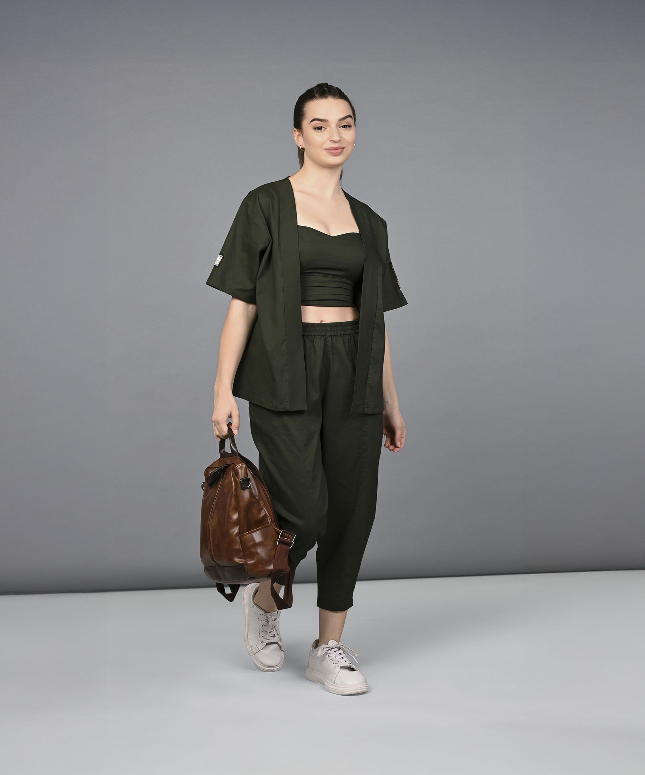 Woman in a Military Green Open Jacket & Yoga Pants with Bustiere set, standing against a plain gray background, carrying a brown backpack.