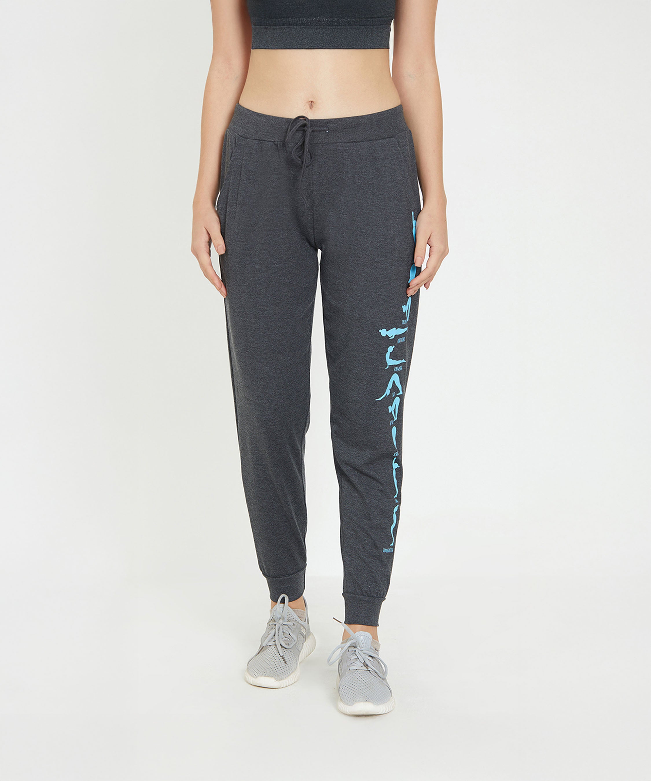 A woman in TUNA LONDON Joggers - Ashtanga Series Grey, paired with a black crop top and gray sneakers, stands confidently against a white background—a chic and comfy look for stylish women.