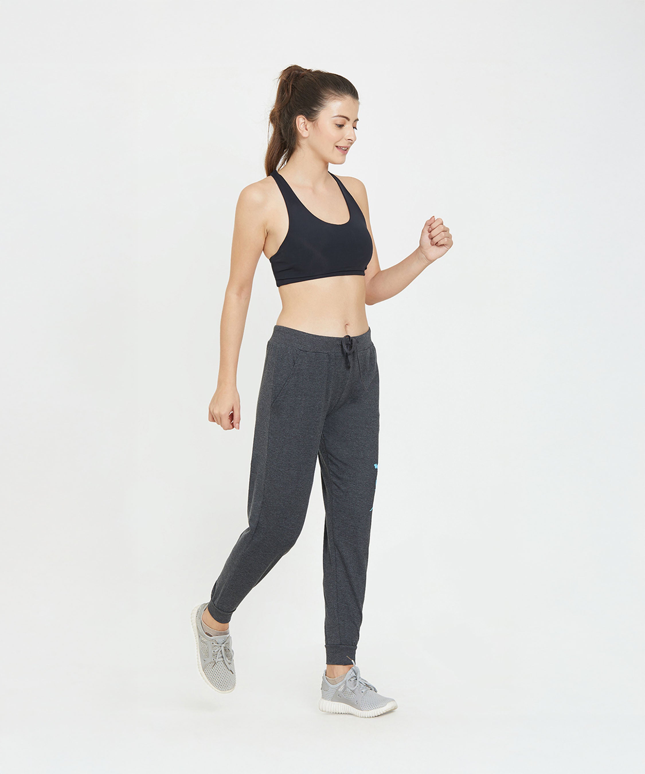 Woman wearing a black sports bra and TUNA LONDON Ashtanga Series Grey joggers, smiling and looking down as she walks against a white background.