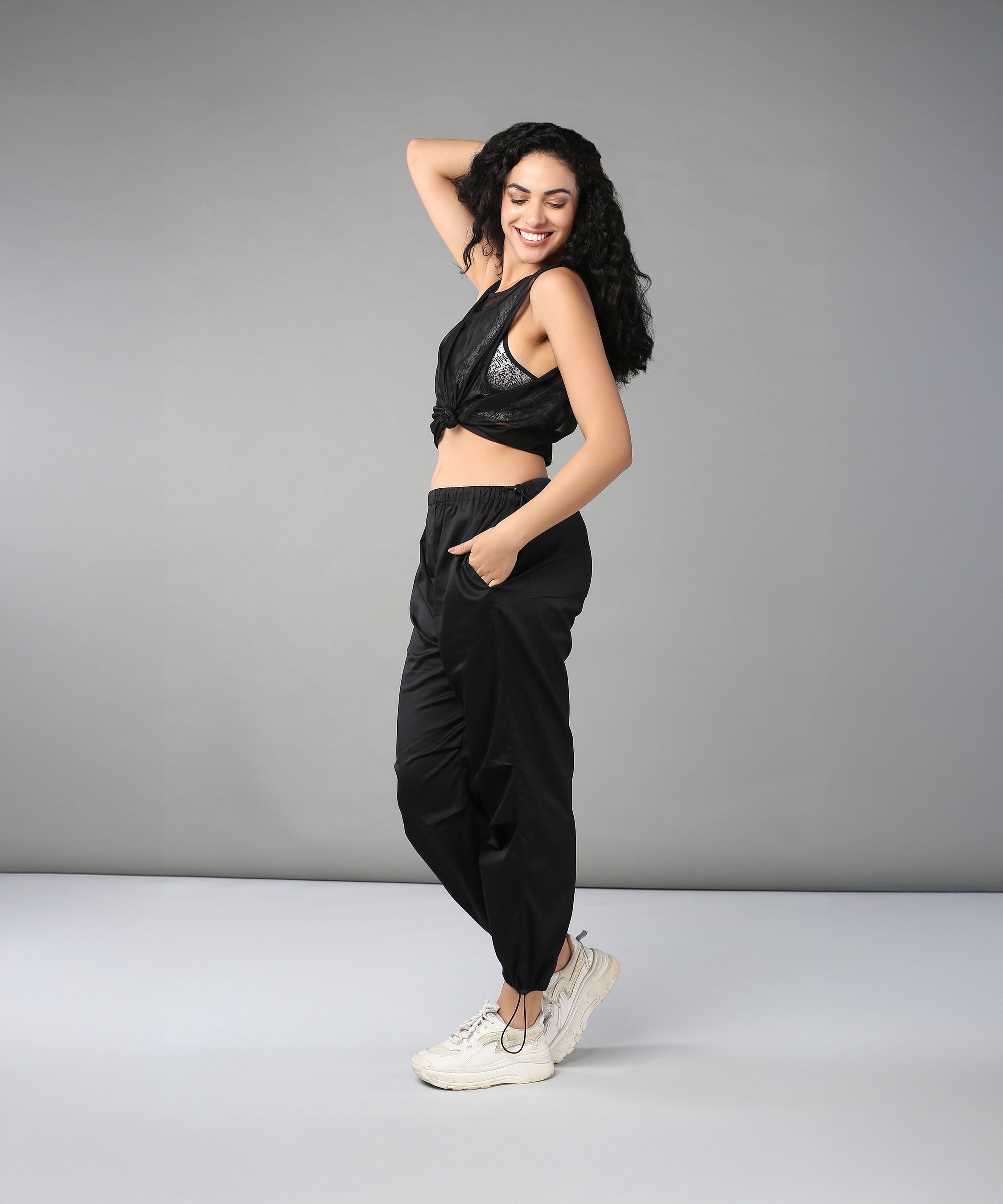 A smiling woman stands sideways against a gray background, posing with her hand on her head while wearing Black Parachute Pants and a crop top.