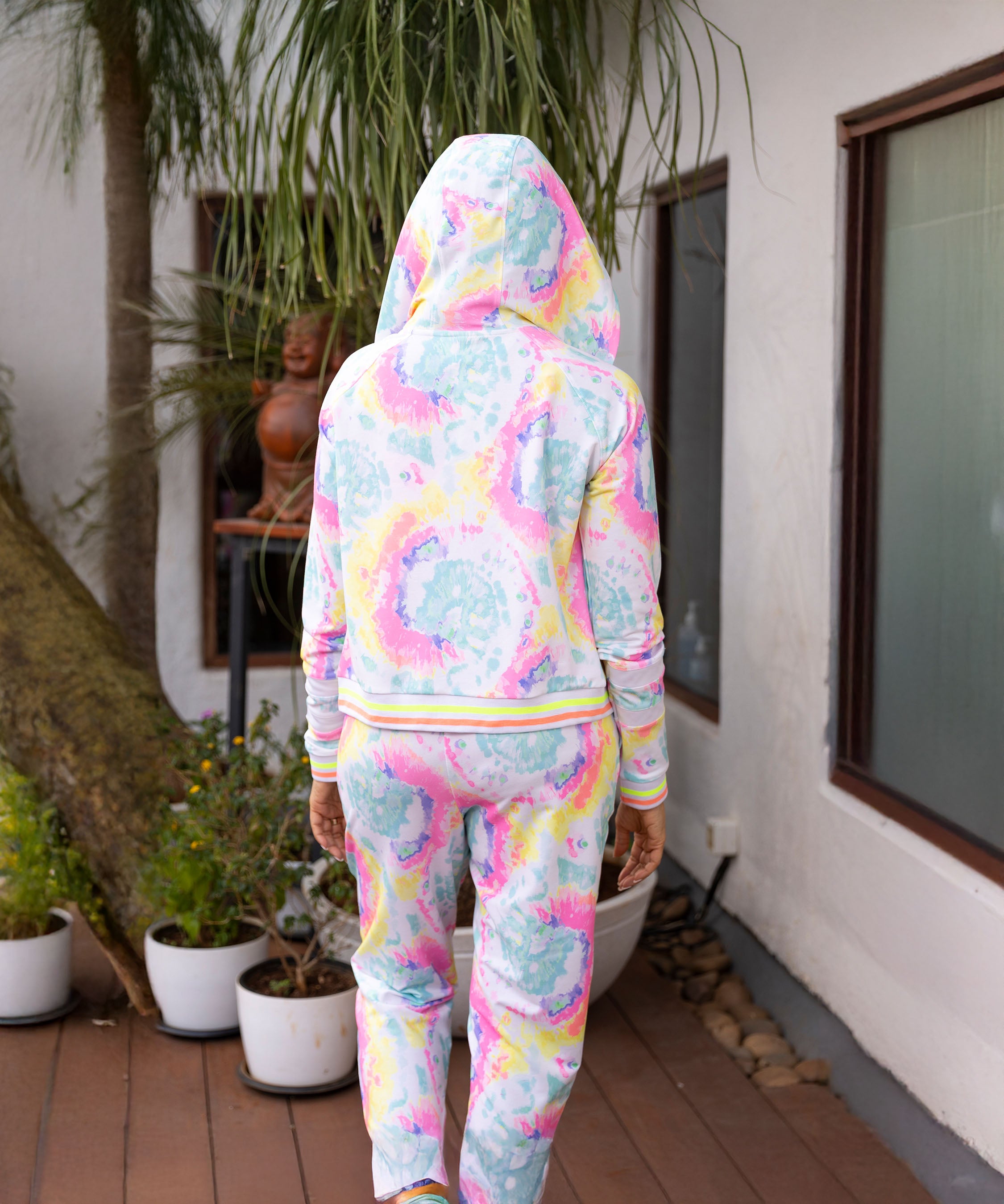 Person in Multicolour Neon Jacket & Joggers Set stands on a wooden deck facing away, with potted plants nearby. This comfy outfit features soft poly blend fabric for a relaxed vibe.