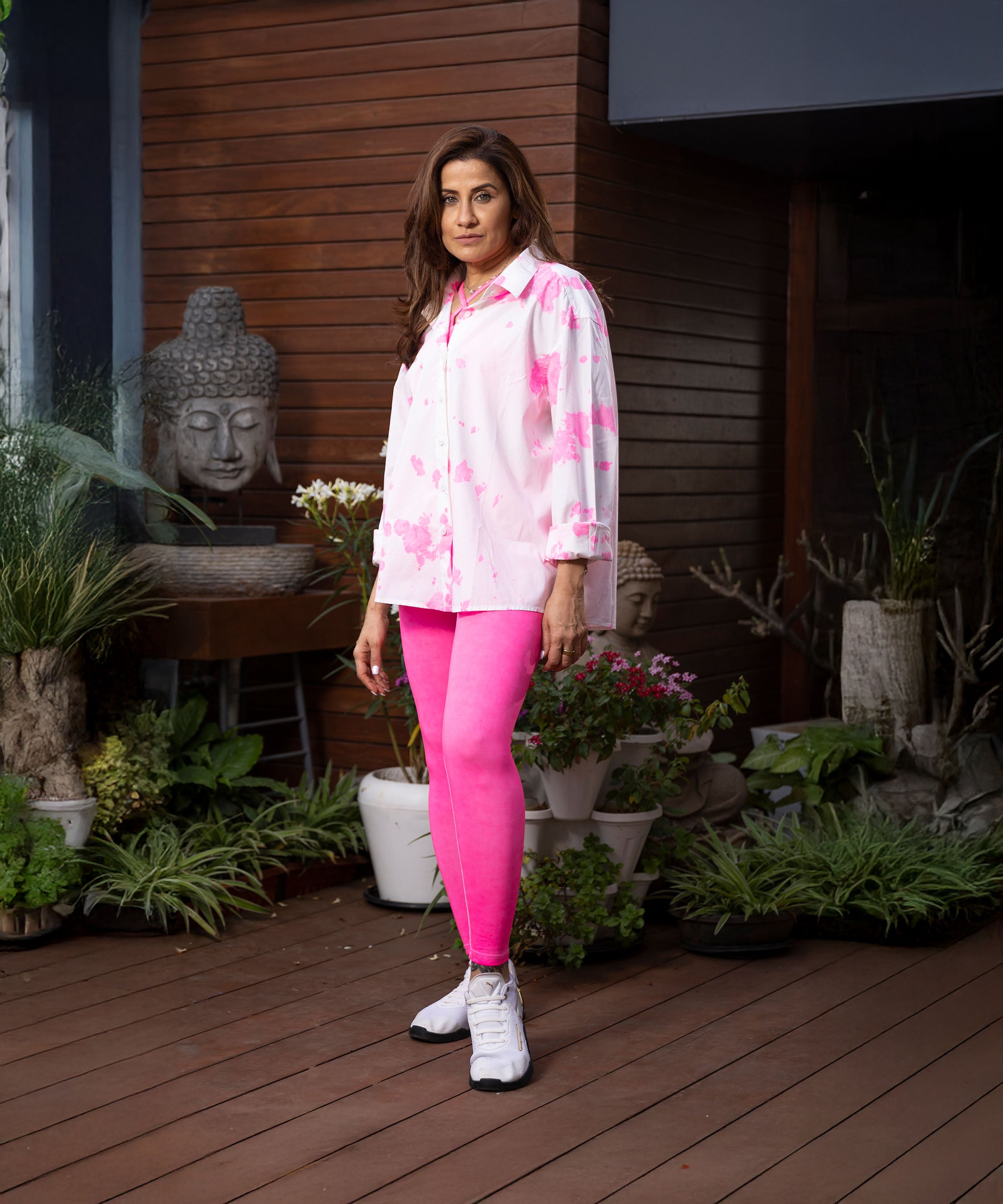 A woman in the Pink Patched Perfection shirt stands on a wooden deck, surrounded by plants and a Buddha statue.