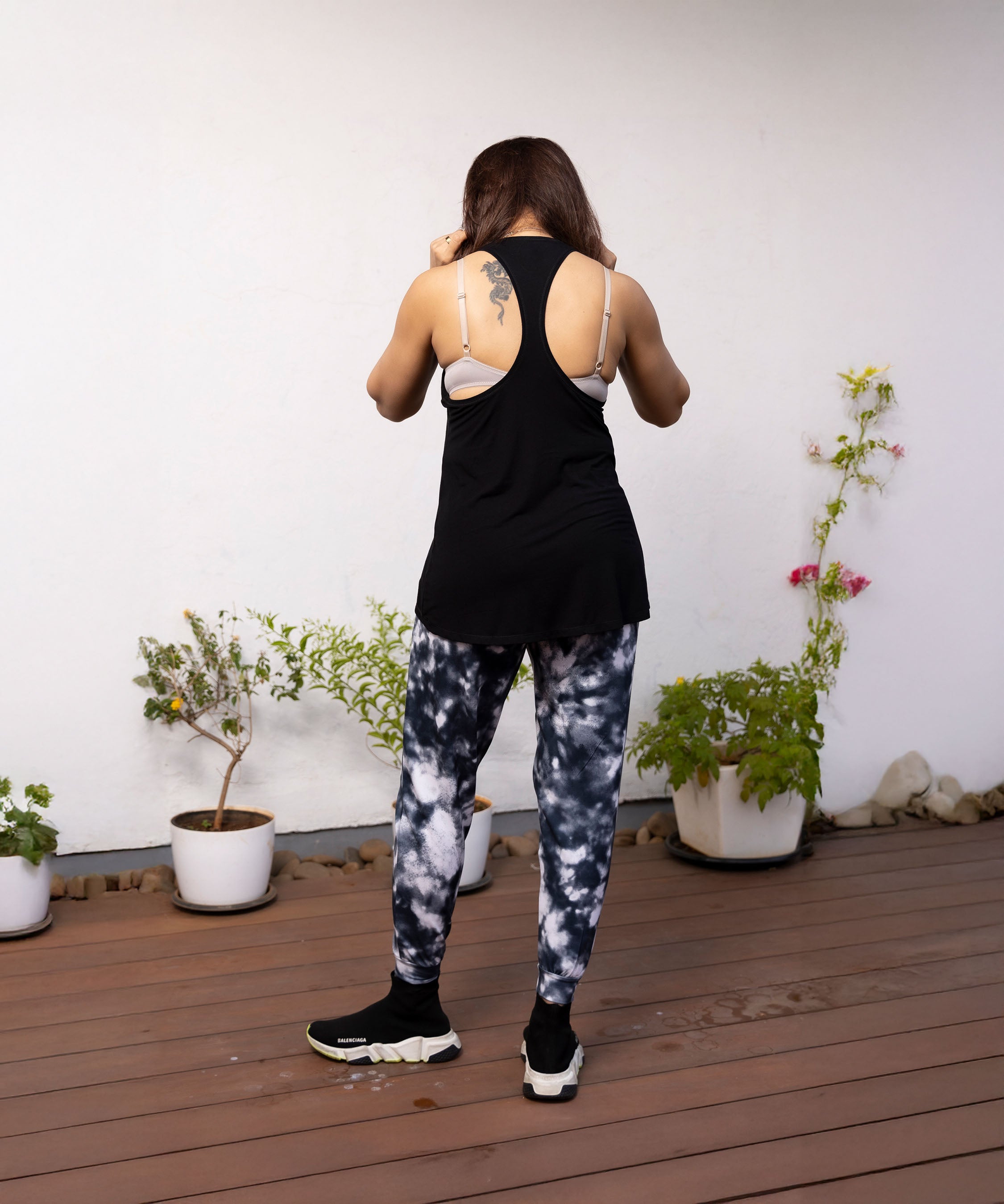 A woman stands on a wooden deck facing away, wearing the 'Adore Your Core' Printed Top with Racer Back near potted plants by a white wall.