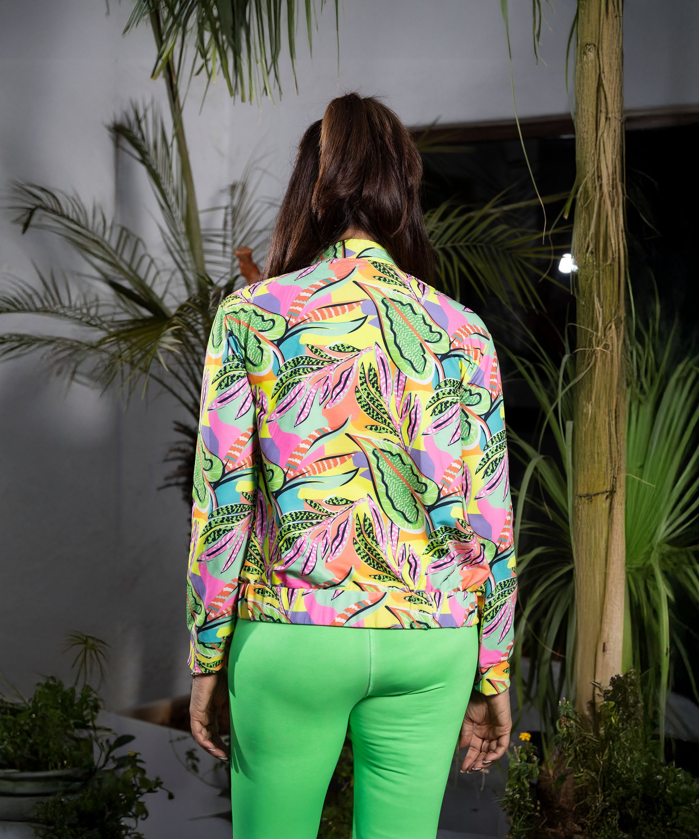 A person with long hair, dressed in a colorful jacket and Neon Green Leggings, stands among lush green plants with their back to the camera.