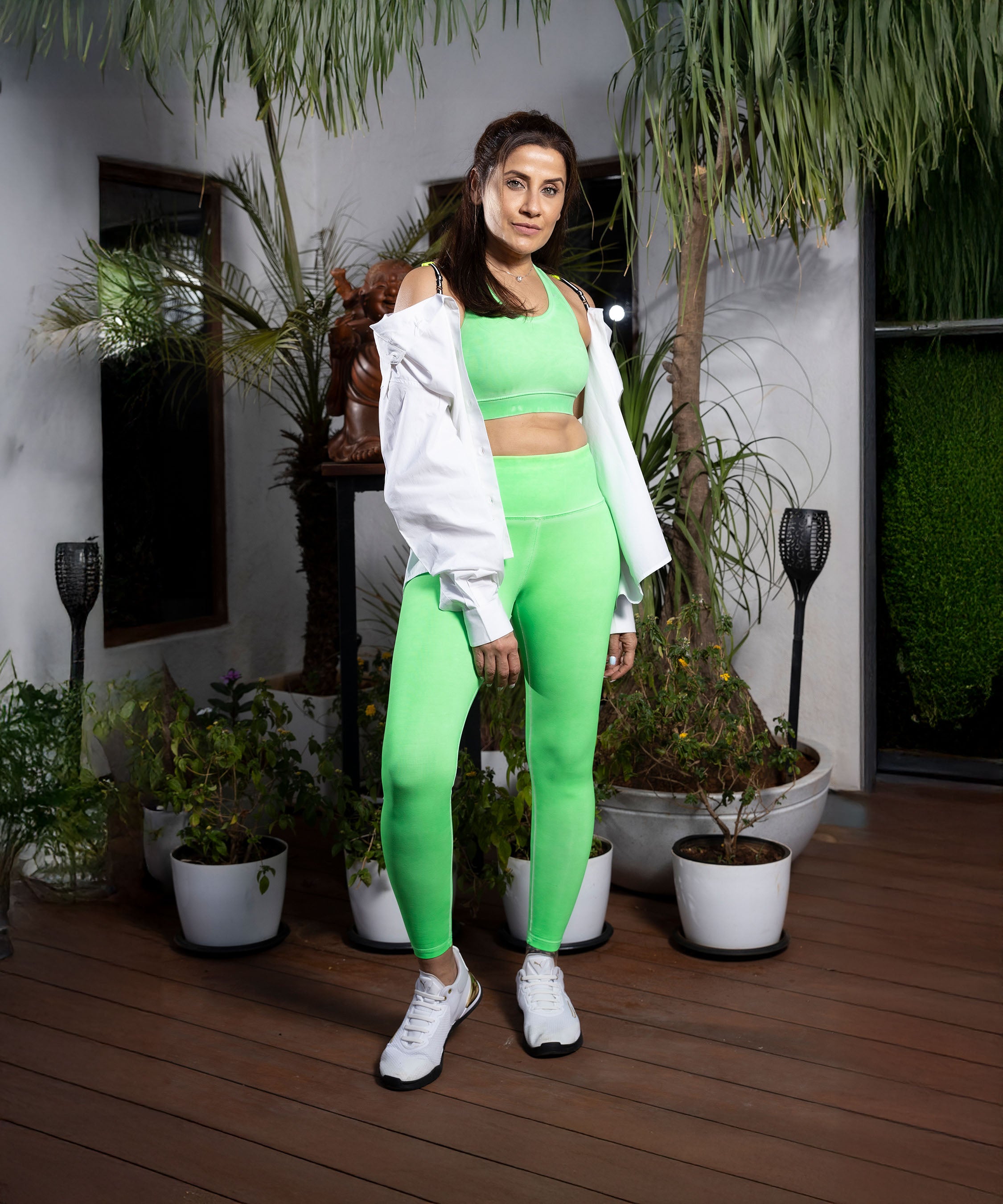 A woman in neon green leggings and a white jacket stands on a patio surrounded by potted plants and greenery.