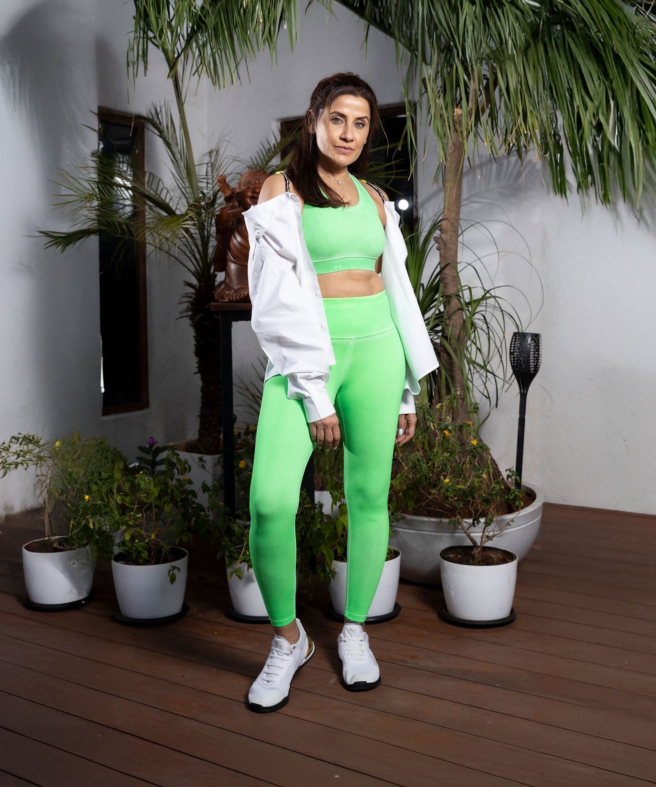 A woman in a comfortable Neon Green Sports Bra and white jacket poses on a deck surrounded by potted plants and palm trees.