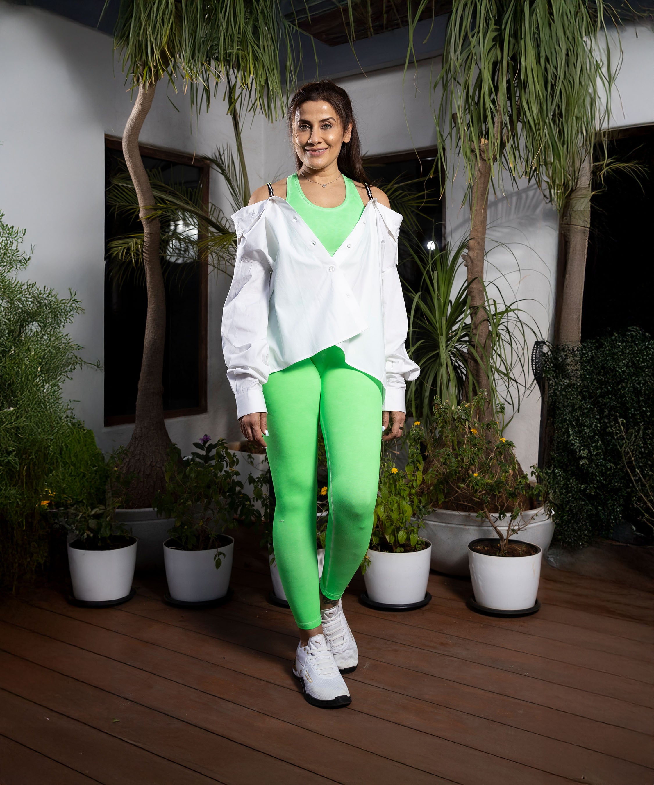 A woman wearing Neon Green Leggings and a white jacket stands on a wooden deck, surrounded by potted plants.