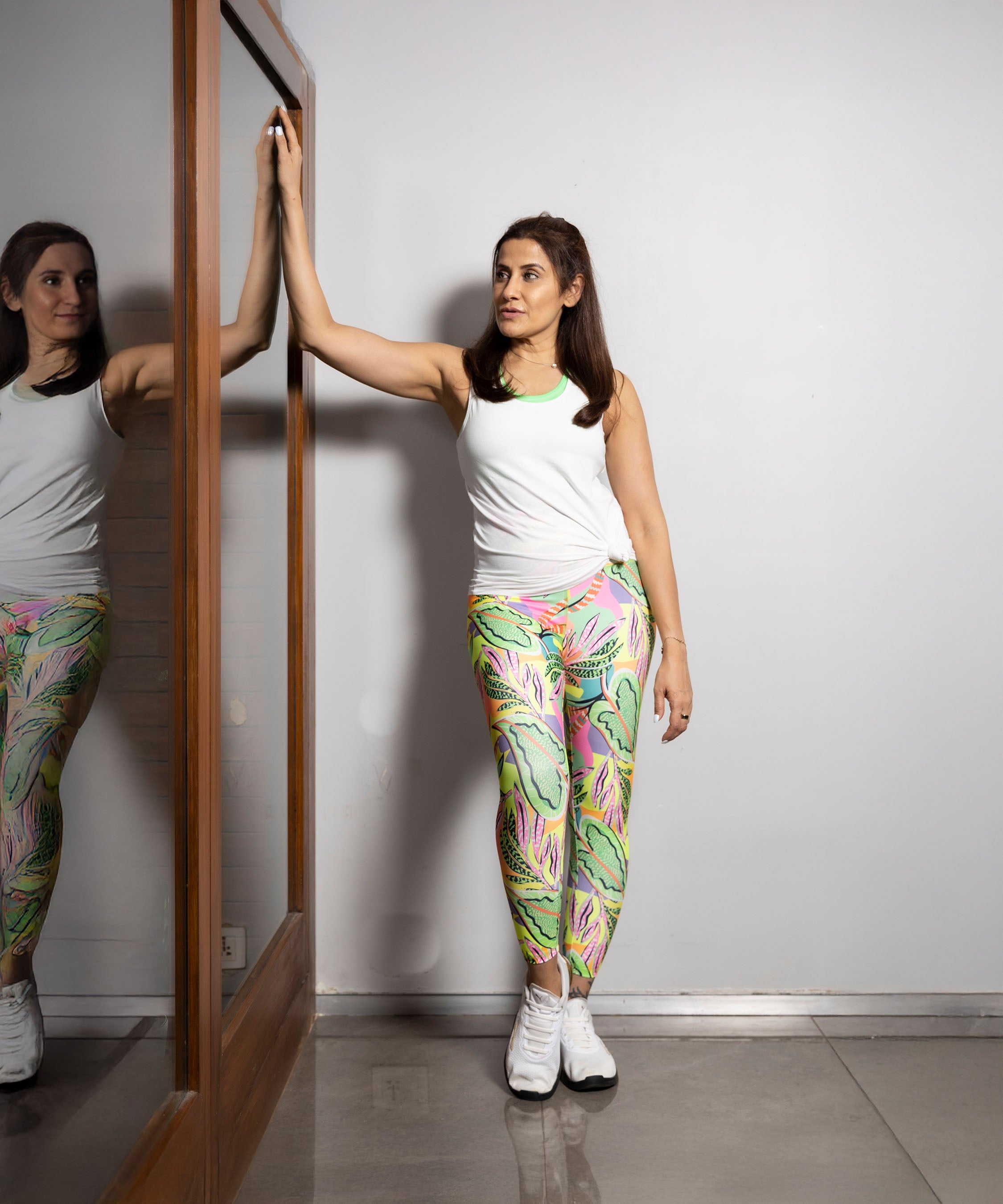 A woman wearing the 'Pilates Has Your Back' Printed Racer Black Top and colorful leggings stands by a mirror, touching it with one hand.