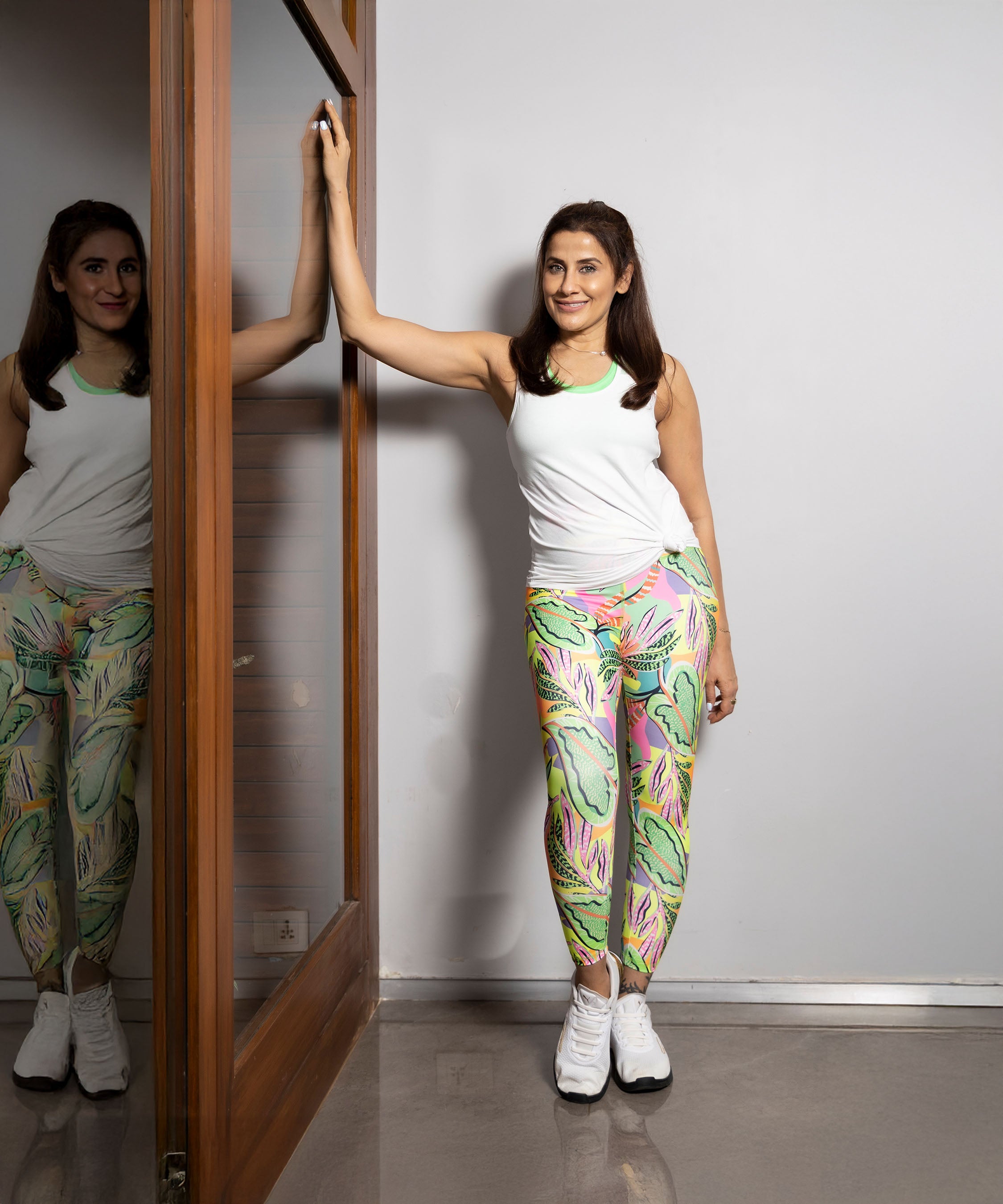 A woman in Multicolor Neon Printed Leggings and a white tank top stretches her arm on a door, smiling indoors. The soft poly blend leggings offer comfort and add a vibrant pop of color to her active routine.