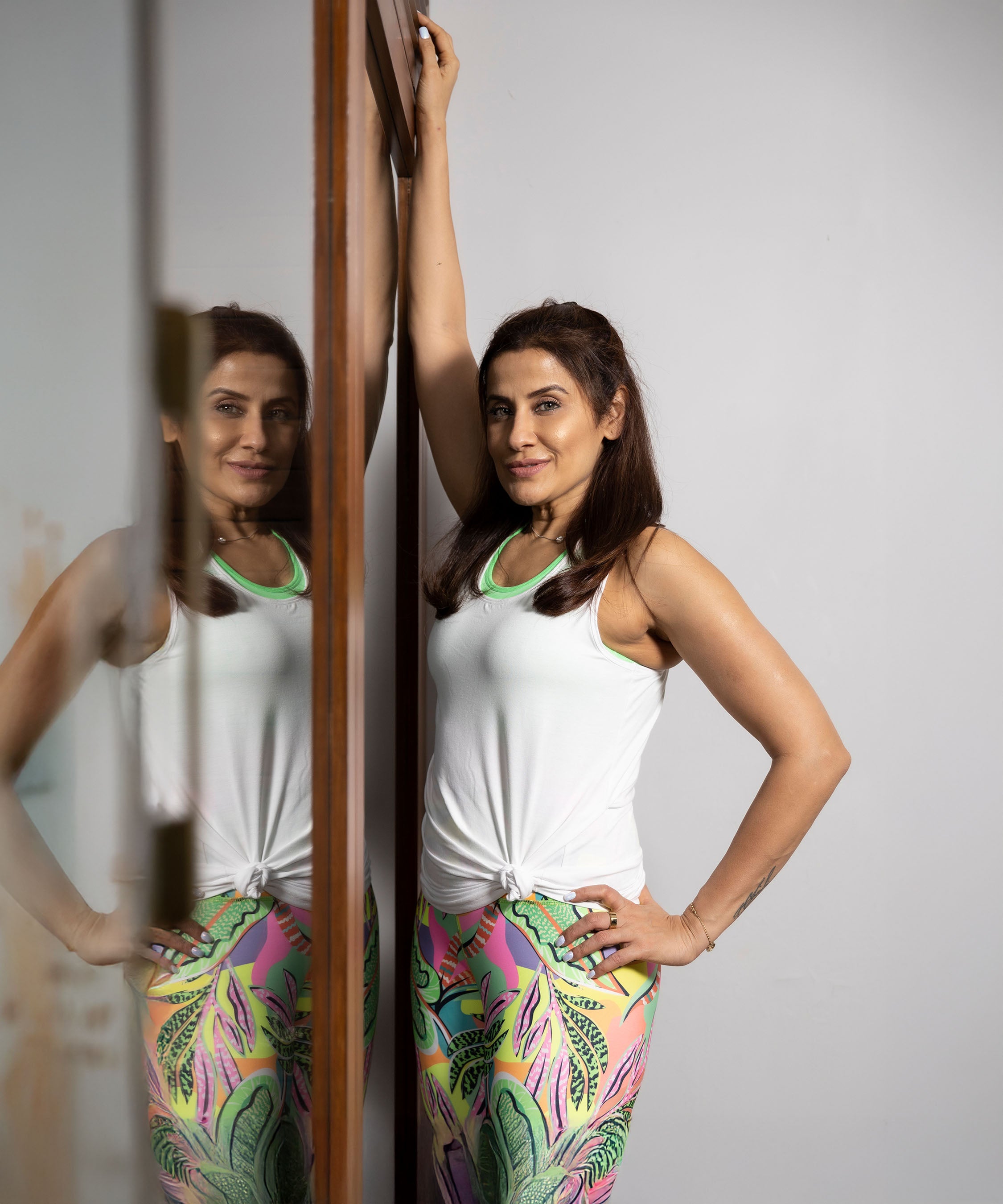 Woman poses confidently by a mirror in the 'Pilates Has Your Back' Printed Racer Black Top and colorful leggings, hand resting on the wall.