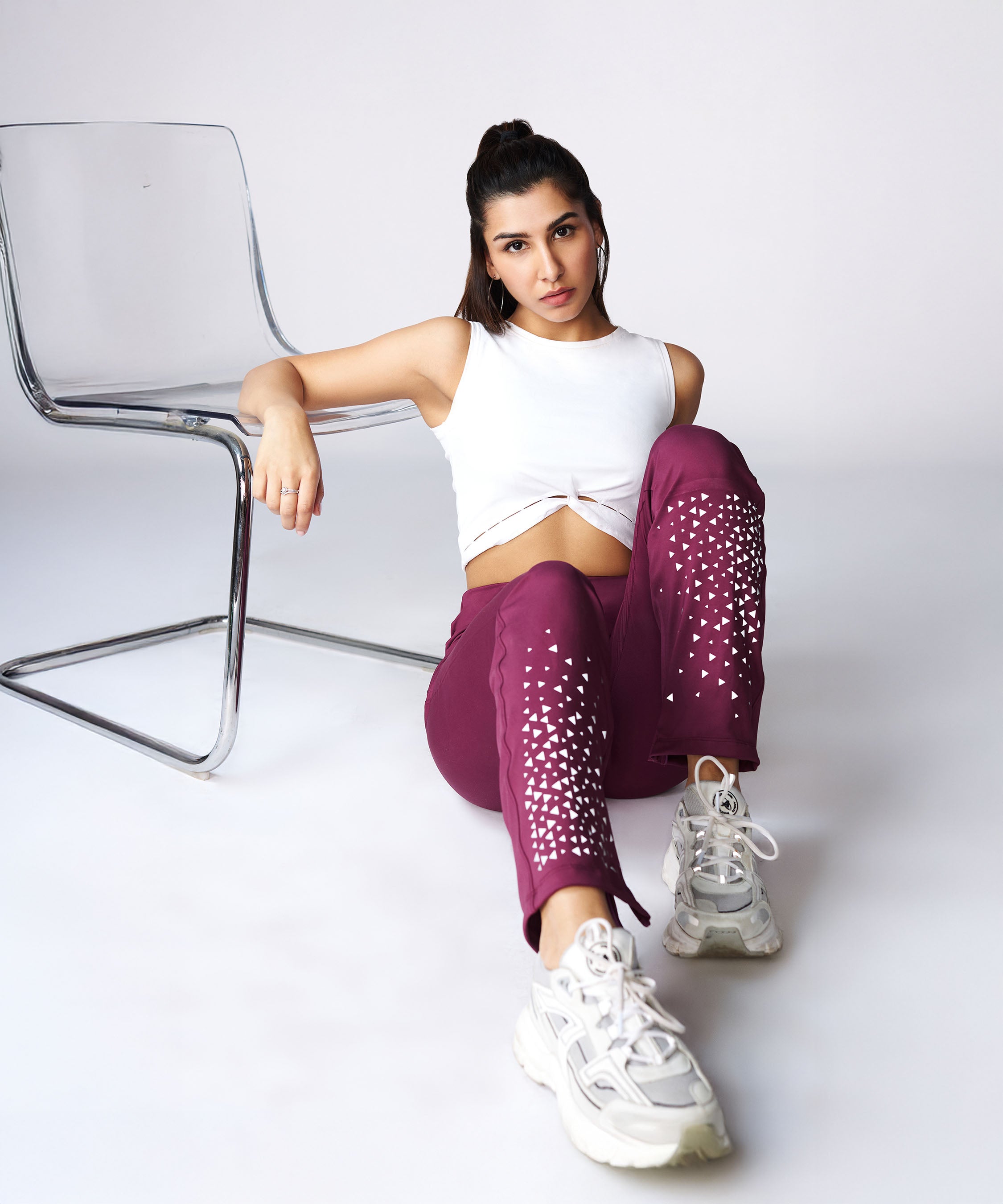 Woman wearing Tuna Active Wine Color Boot Cut Pants and a white crop top sits on the floor beside a clear chair, looking at the camera.