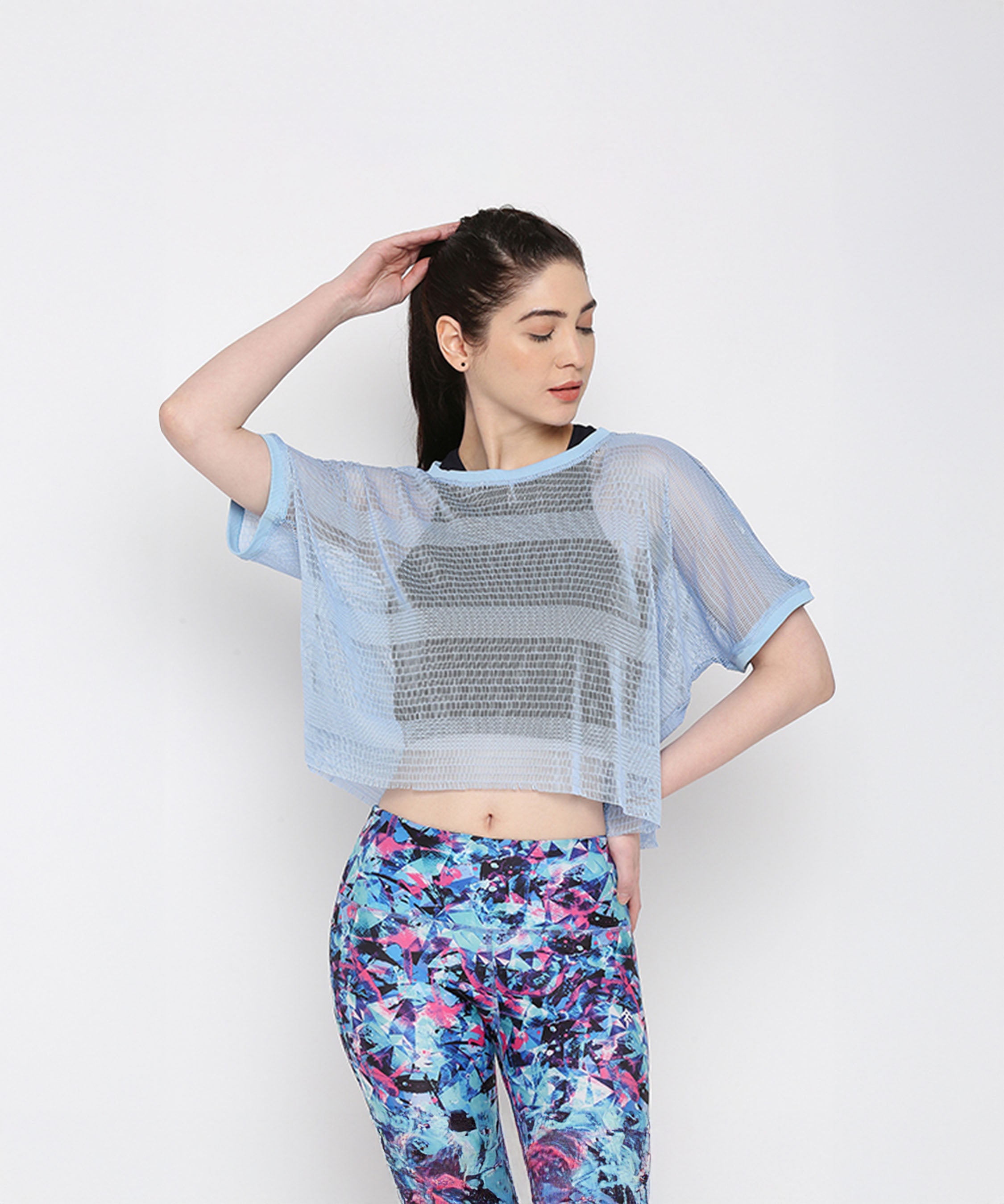 A woman models the Blue Netted Top layered over a black sports bra and vibrant floral leggings, posing against a plain background for a chic, sporty vibe.