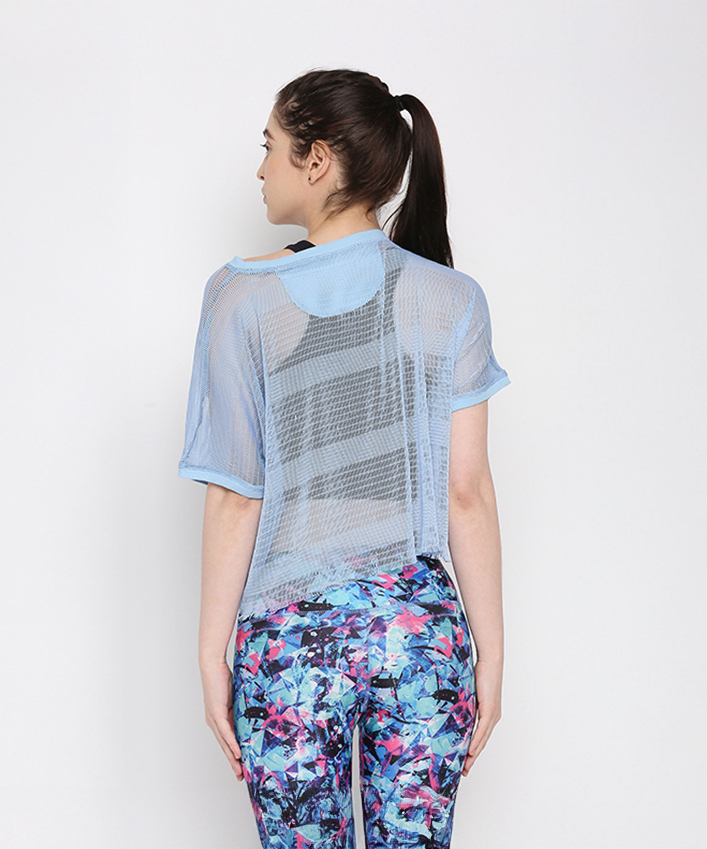 A woman faces away, wearing the Blue Netted Top layered over a striped shirt and stylish, colorful patterned leggings.