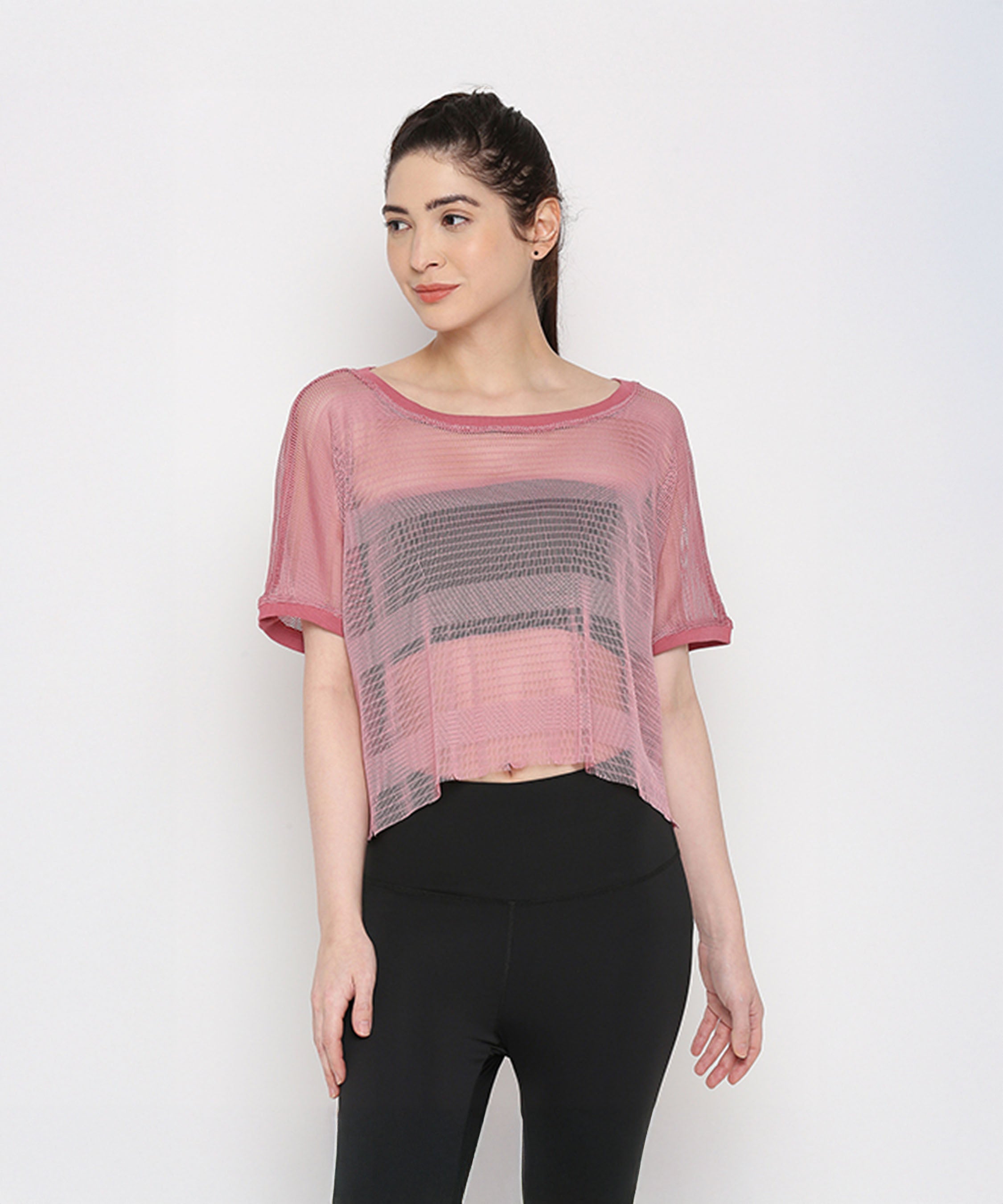 A woman showcases the Pink Netted Top, worn sheer over a black sports bra and paired with stylish leggings, creating a trendy sporty look against a white background.