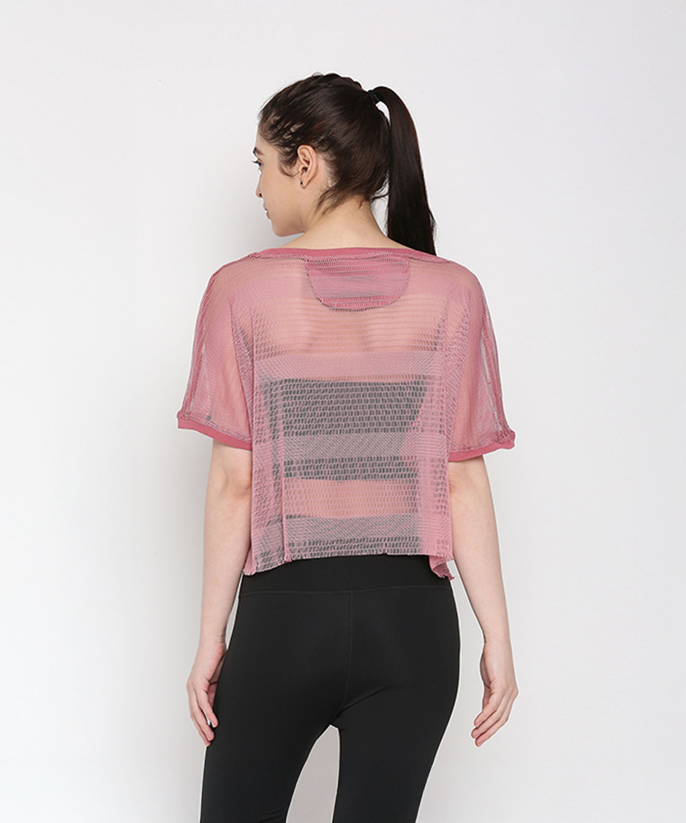 A woman with her hair in a ponytail faces away, wearing the Pink Netted Top in sheer pink over a black sports bra and paired with stylish leggings for a sporty look.