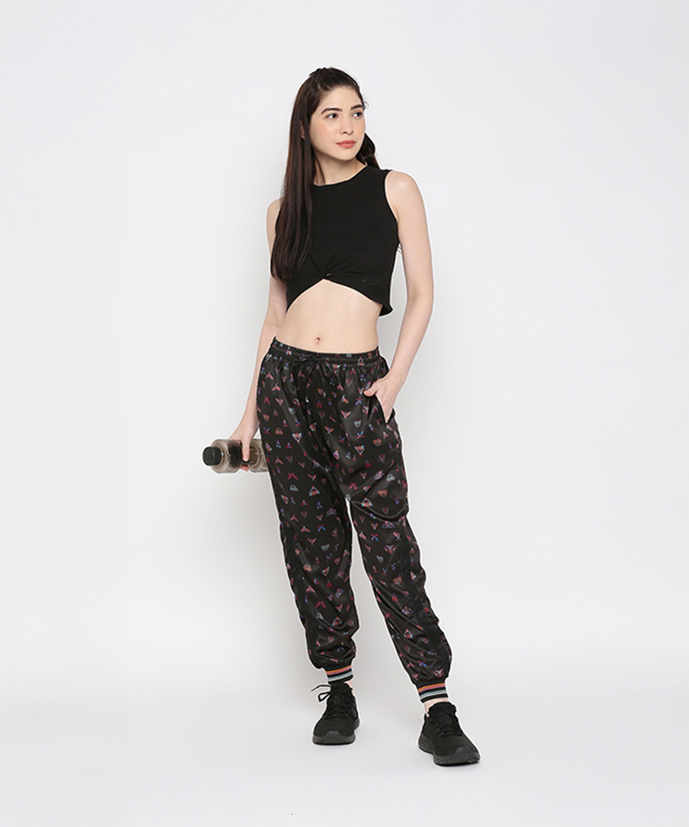 A woman poses against a white background wearing a Black Twisted Crop Top, printed joggers, and sneakers while holding a water bottle.