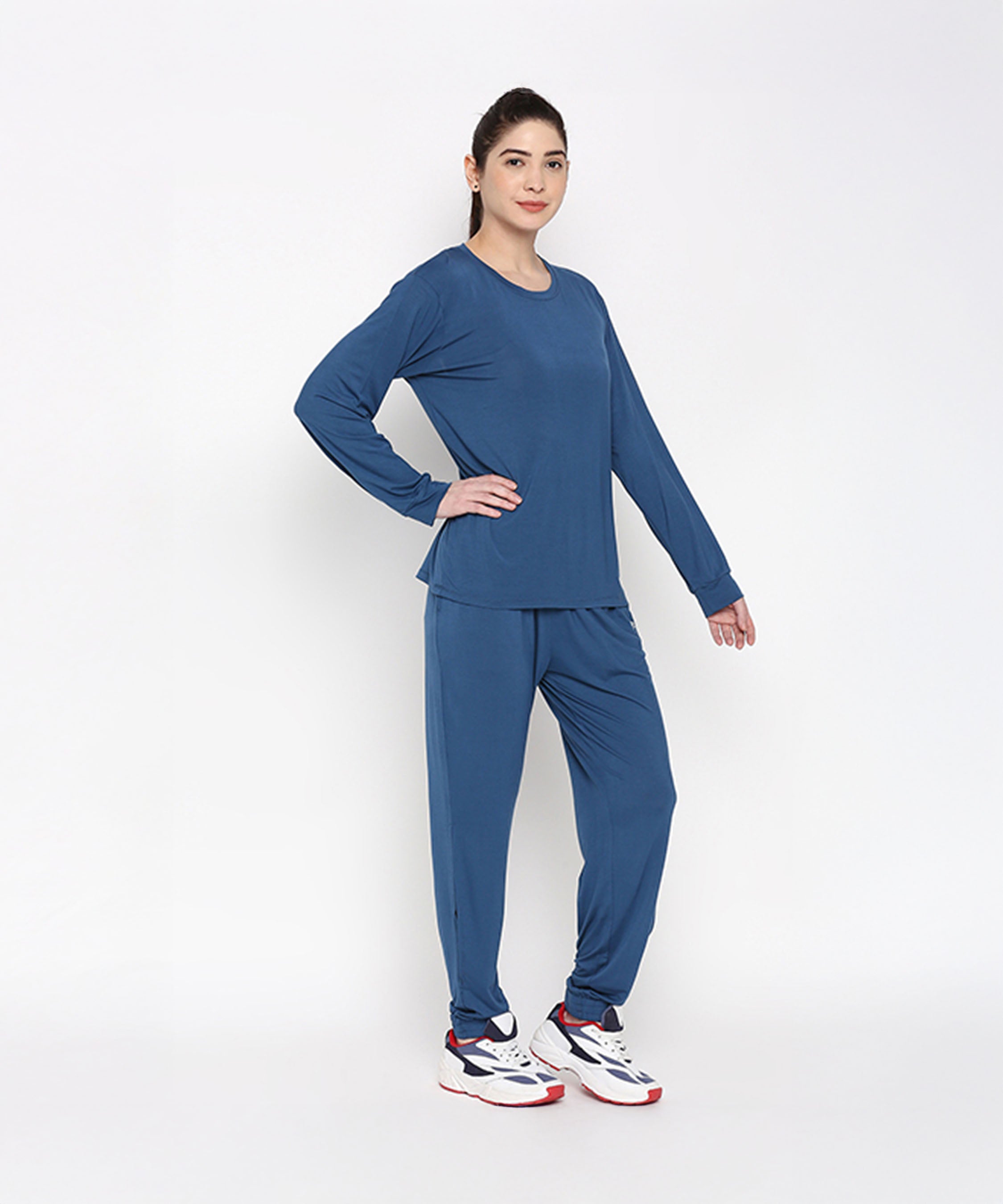 A woman models the Atlantic Blue Active Duo—cotton modal joggers paired with a blue racerback top—and white sneakers, standing against a plain white background.