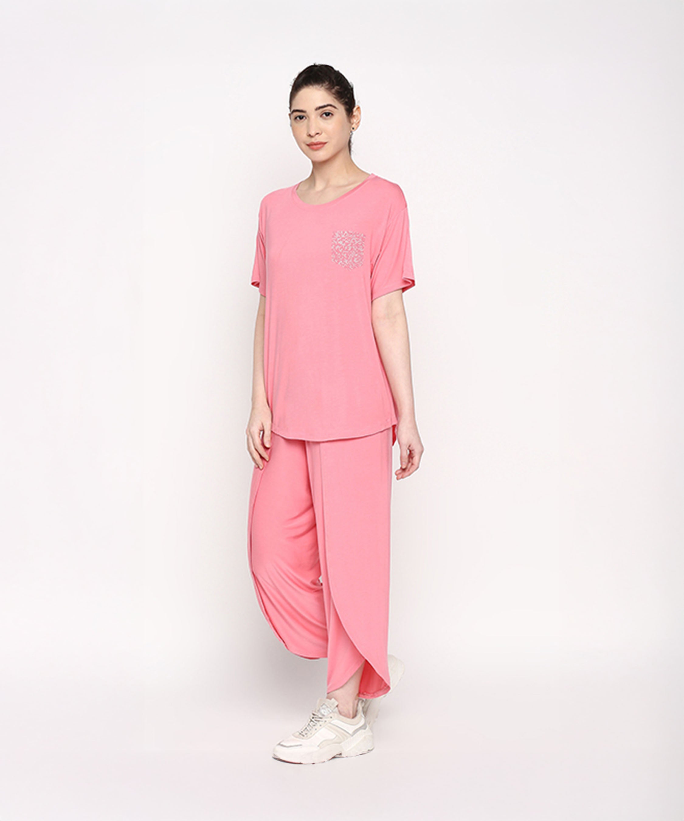 A woman wears the Coral Blissful Tulip Set—coral pink top and matching tulip pants in soft cotton modal—paired with white sneakers, standing against a white background.