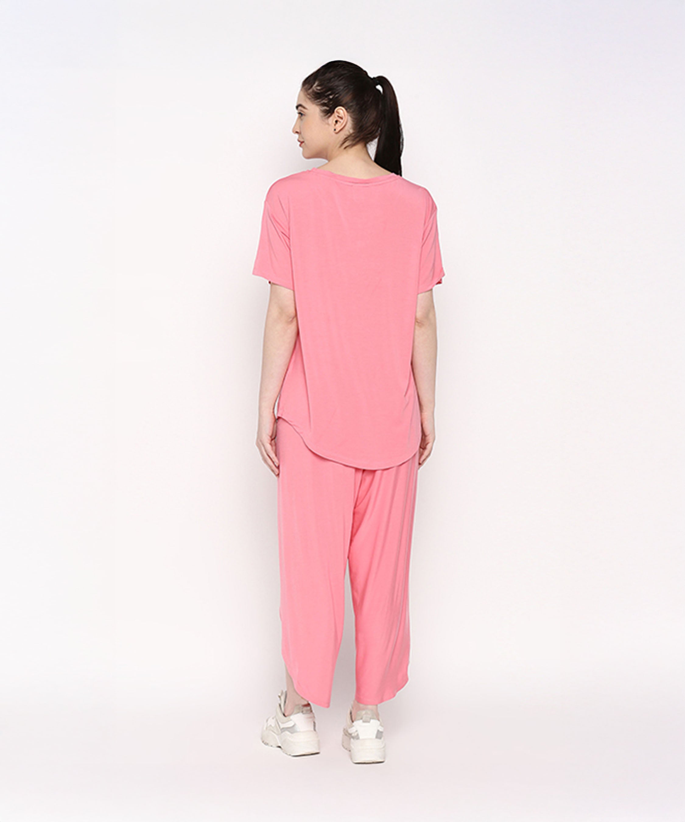 A woman stands facing away against a plain white background, wearing the Coral Blissful Tulip Set— a cotton modal coral pink top and tulip pant loungewear ensemble—paired with white sneakers.