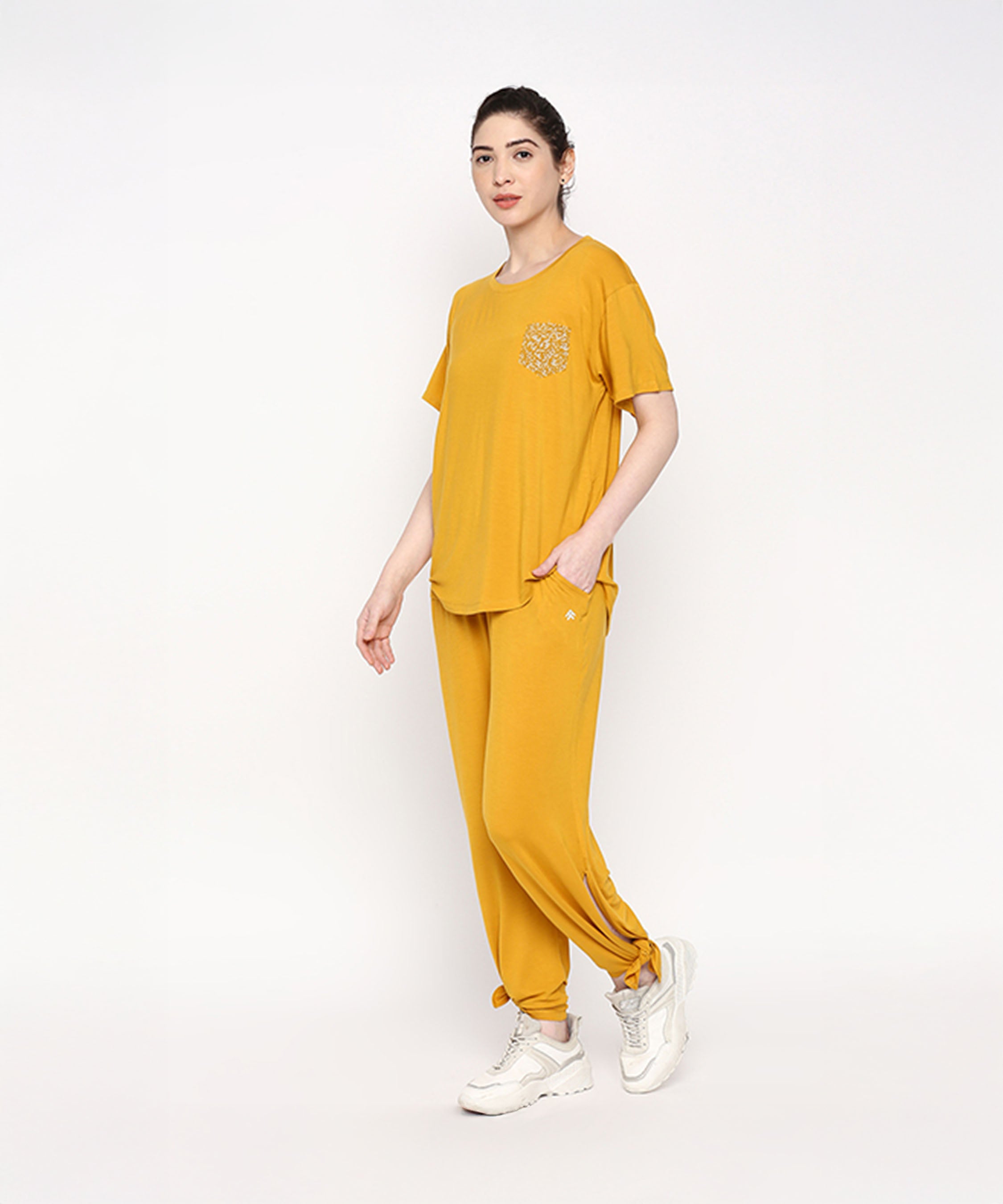 A woman wears the Mustard Chic Set—featuring a mustard yellow top and cotton modal trackpants with a soft elasticated waistband—paired with white sneakers, standing against a plain white background.