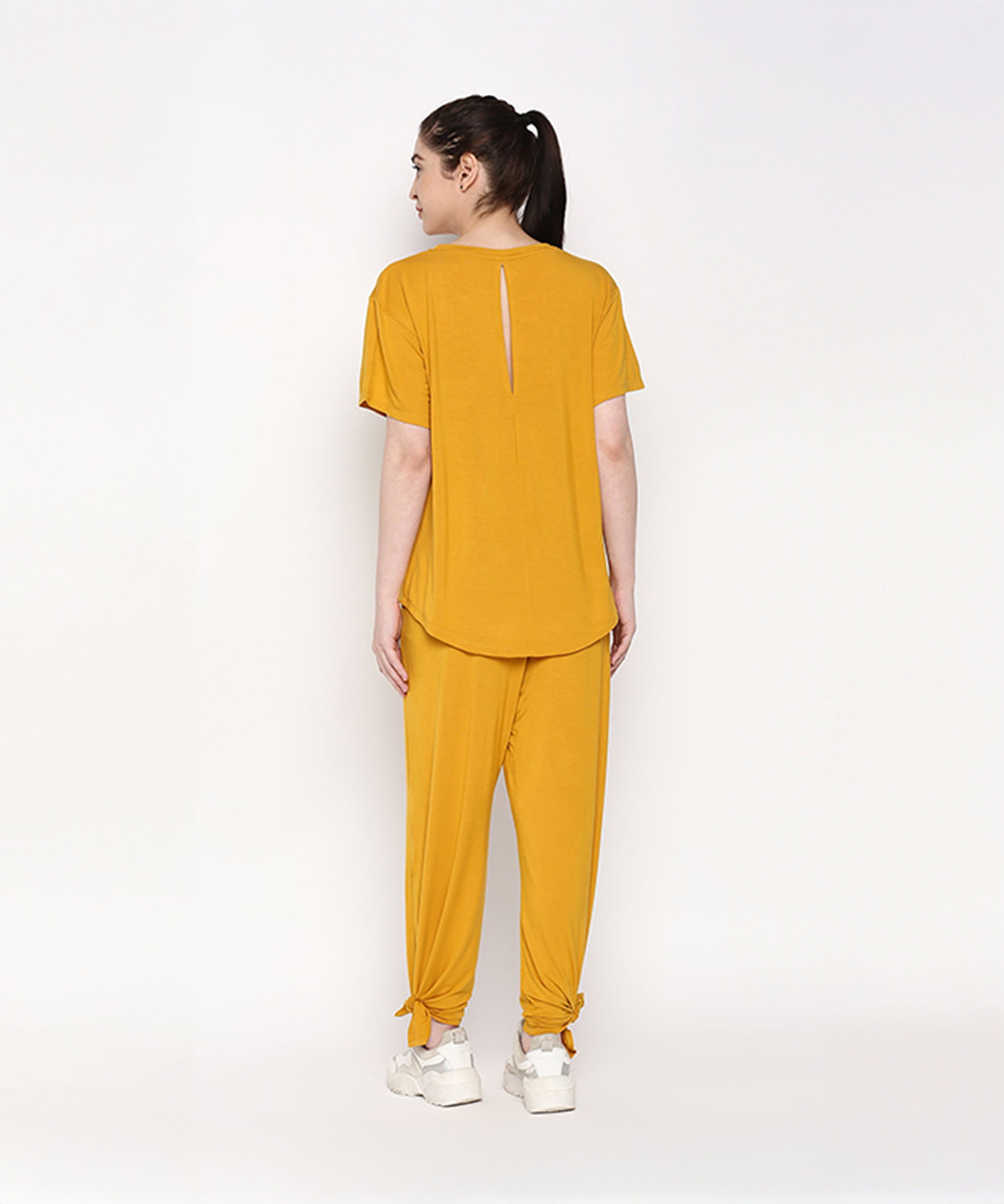 A woman models the Mustard Chic Set—featuring a mustard yellow top and cotton modal trackpant with side knots and a soft elasticated waistband—posed against a white background, facing away.