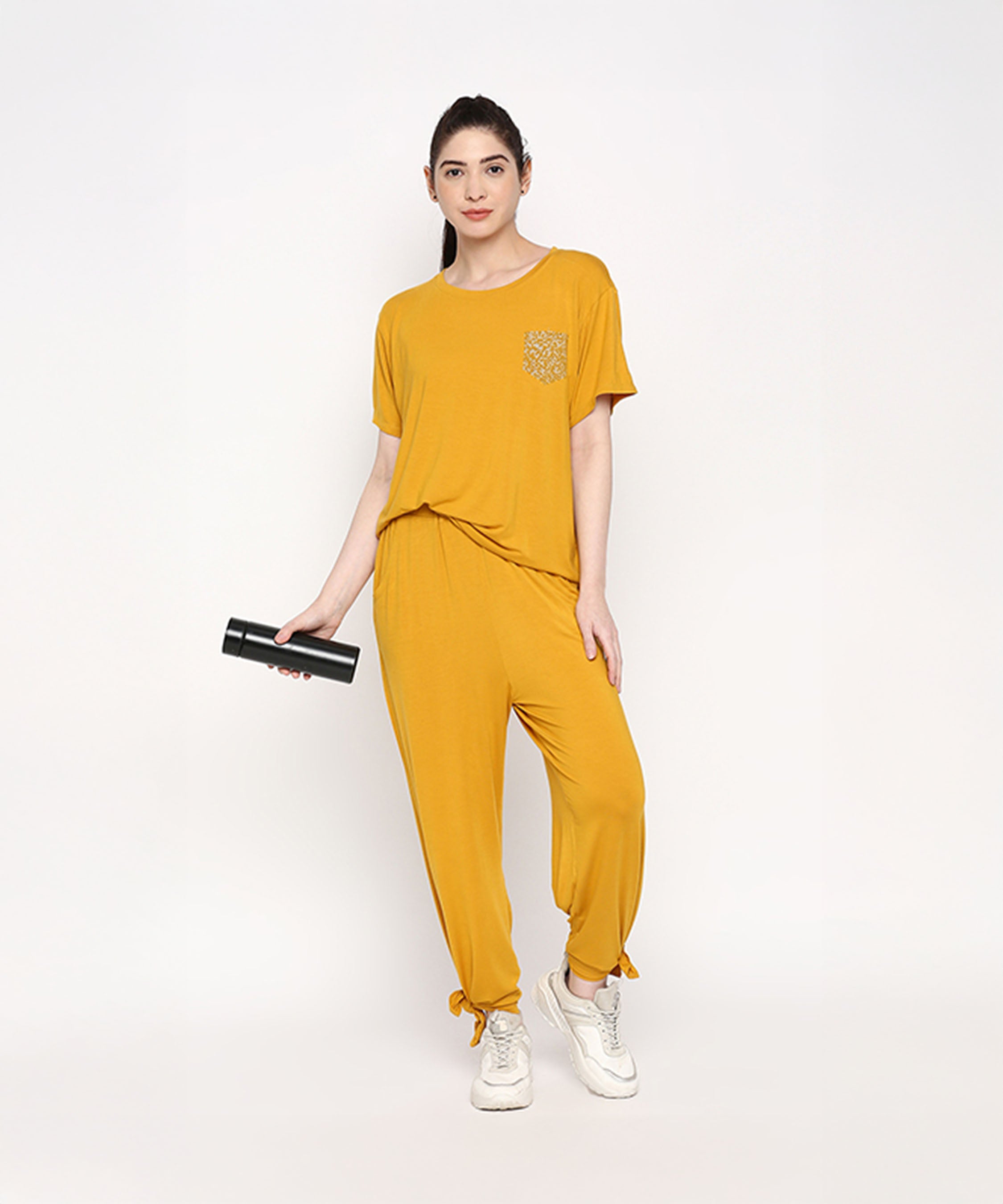 Woman wearing the Mustard Chic Set—mustard yellow top and cotton modal trackpants with a soft elasticated waist—paired with white sneakers, holding a black bottle against a white background.