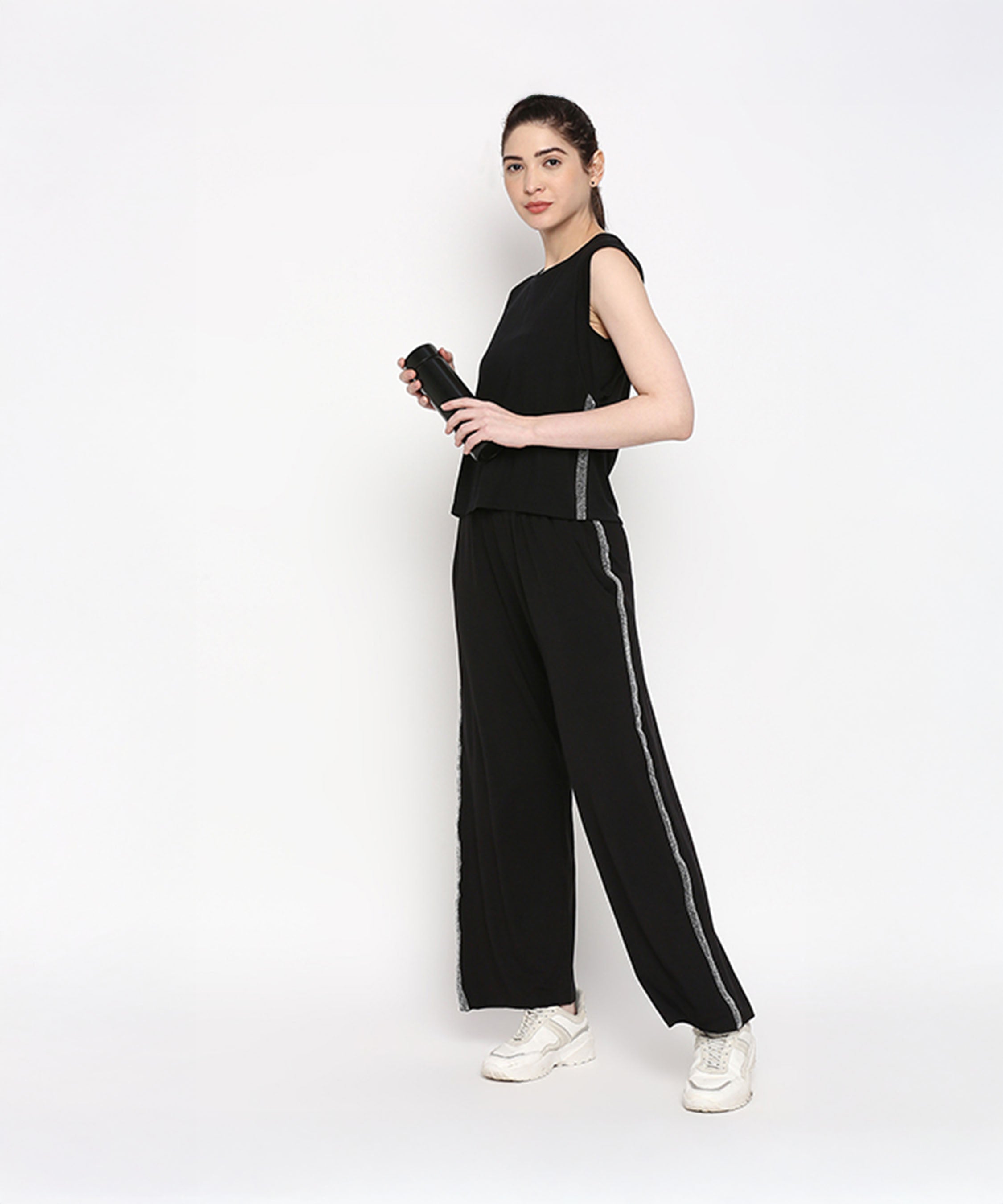 Woman wearing the Comfy Black Set with Side Sparkle, featuring a black sleeveless top and loose-fit track pant, stands against a white background holding a black bottle.