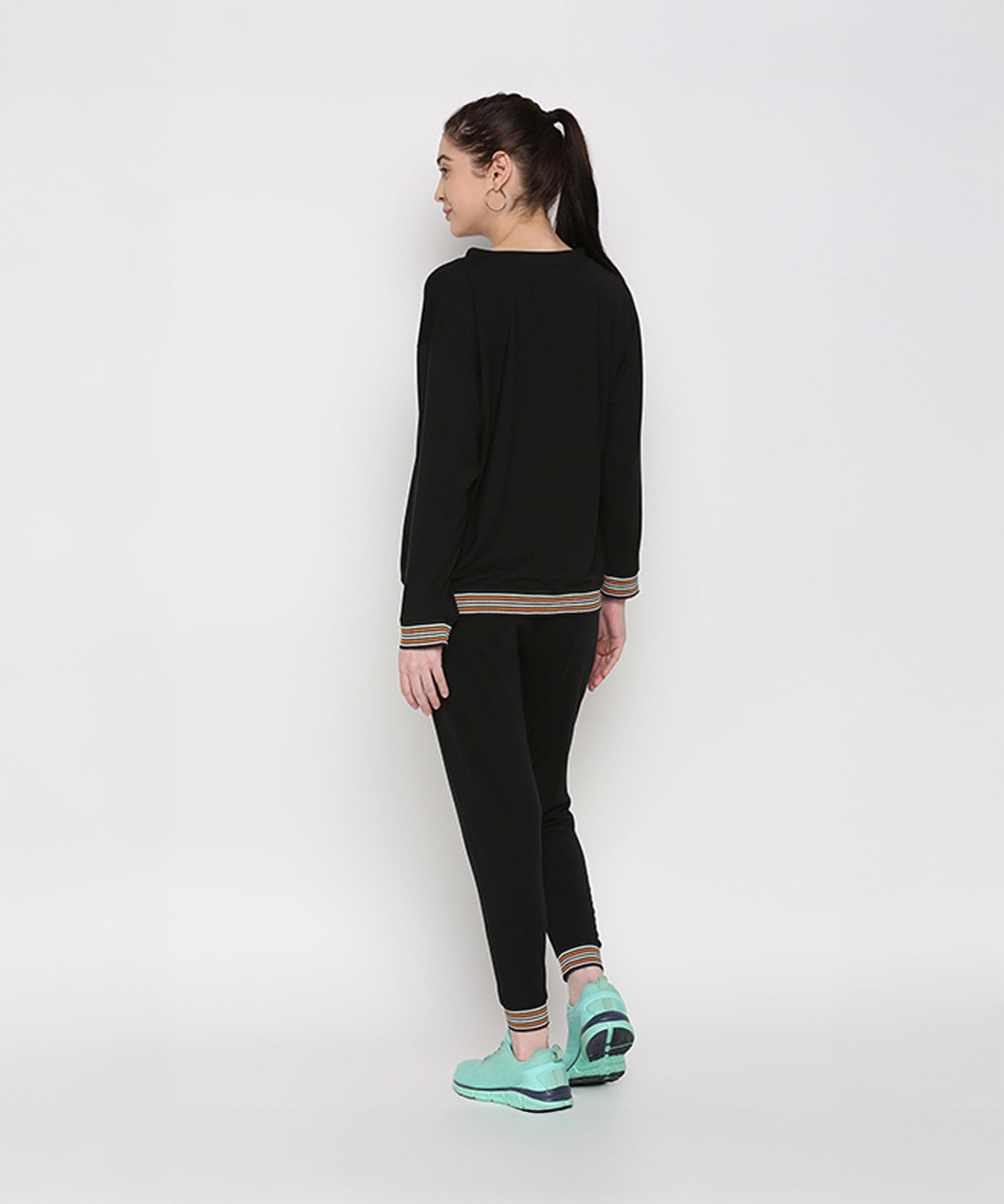 A woman models the Versatile Black Ensemble, featuring a black neck cut out top and cotton modal joggers with teal sneakers, standing sideways against a white background.