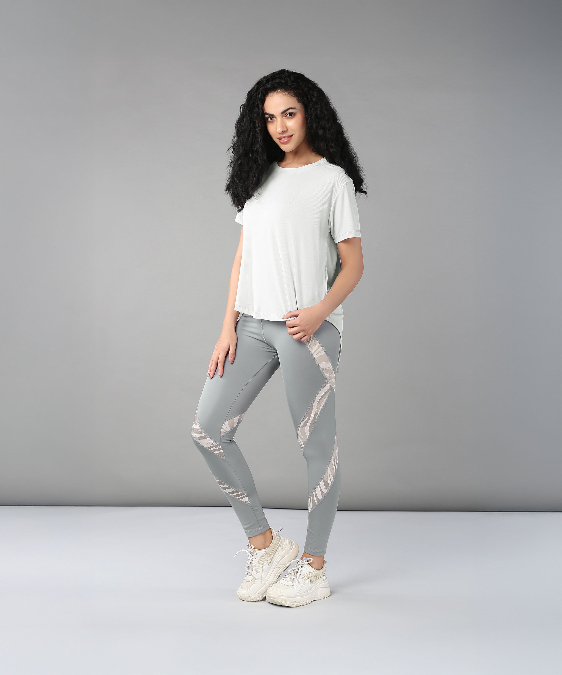 Woman with long curly hair wears the Grey Top with Patched Legging, made from super soft, moisture-wicking fabric, paired with sneakers and poses against a plain gray background.