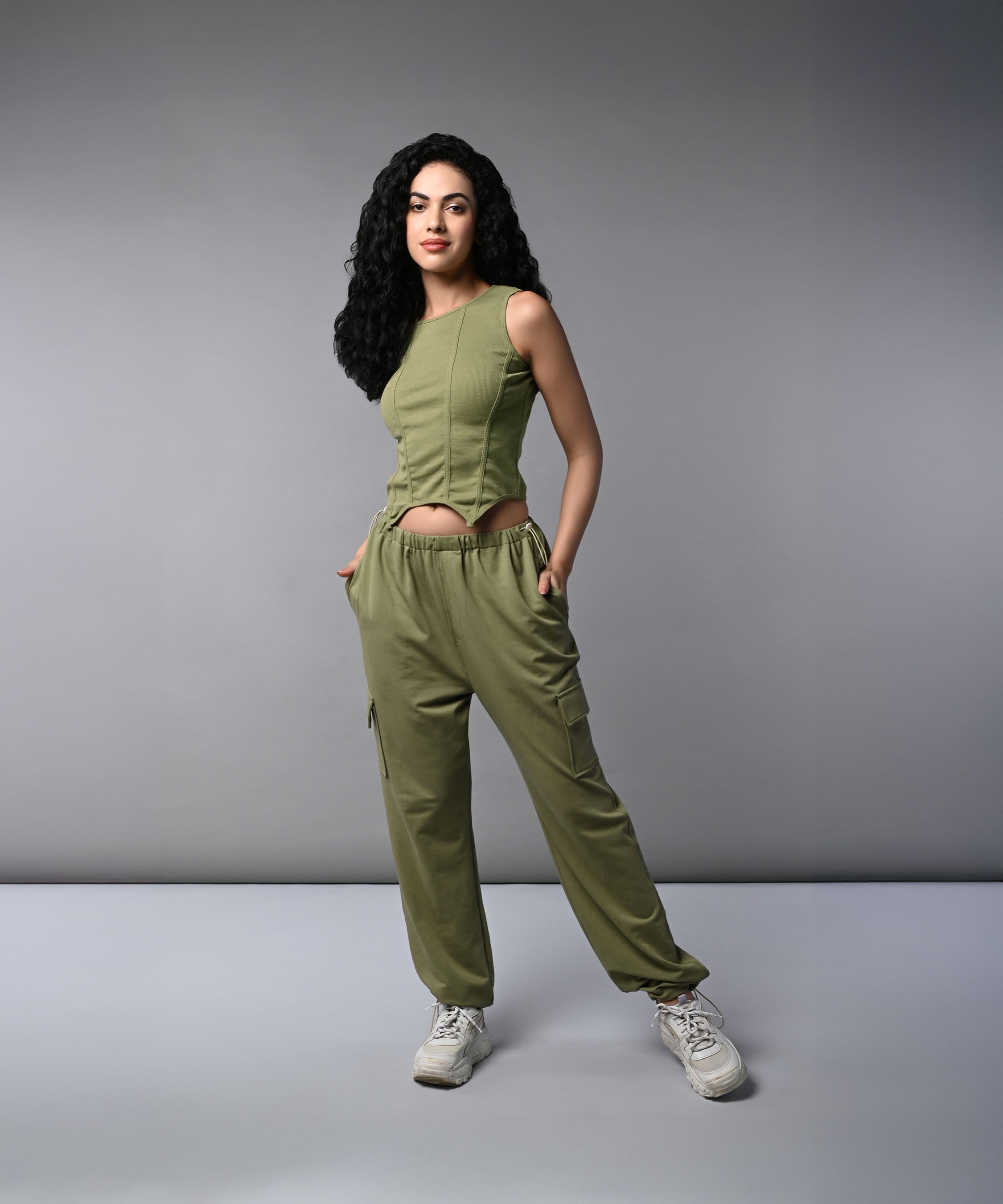 A woman with long dark hair models the Olive Corset Crop - Top with Parachute Pants, standing hands-in-pockets against a gray background.