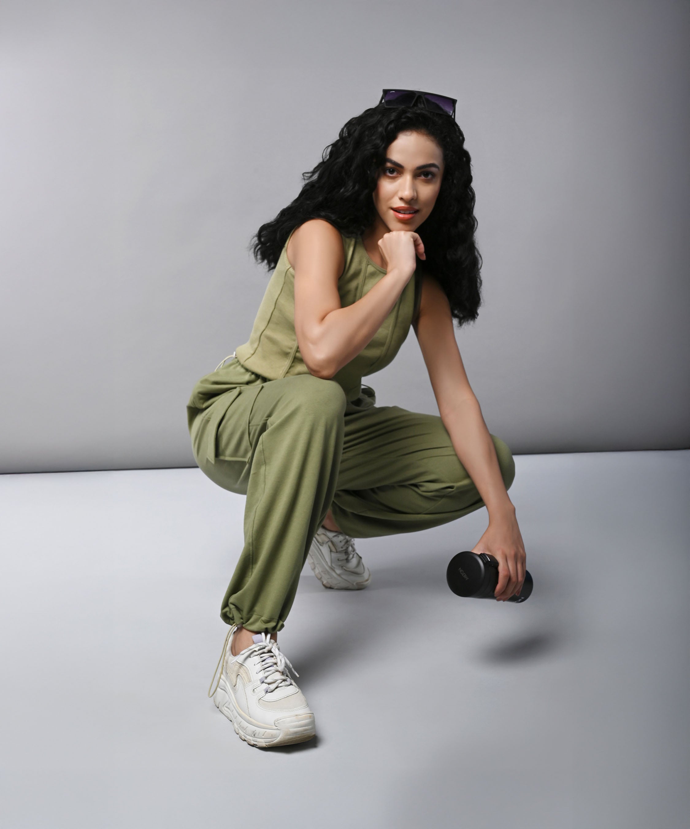 Woman wears the Olive Corset Crop - Top with Parachute Pants co-ord, crouching and holding a dumbbell against a plain gray background.