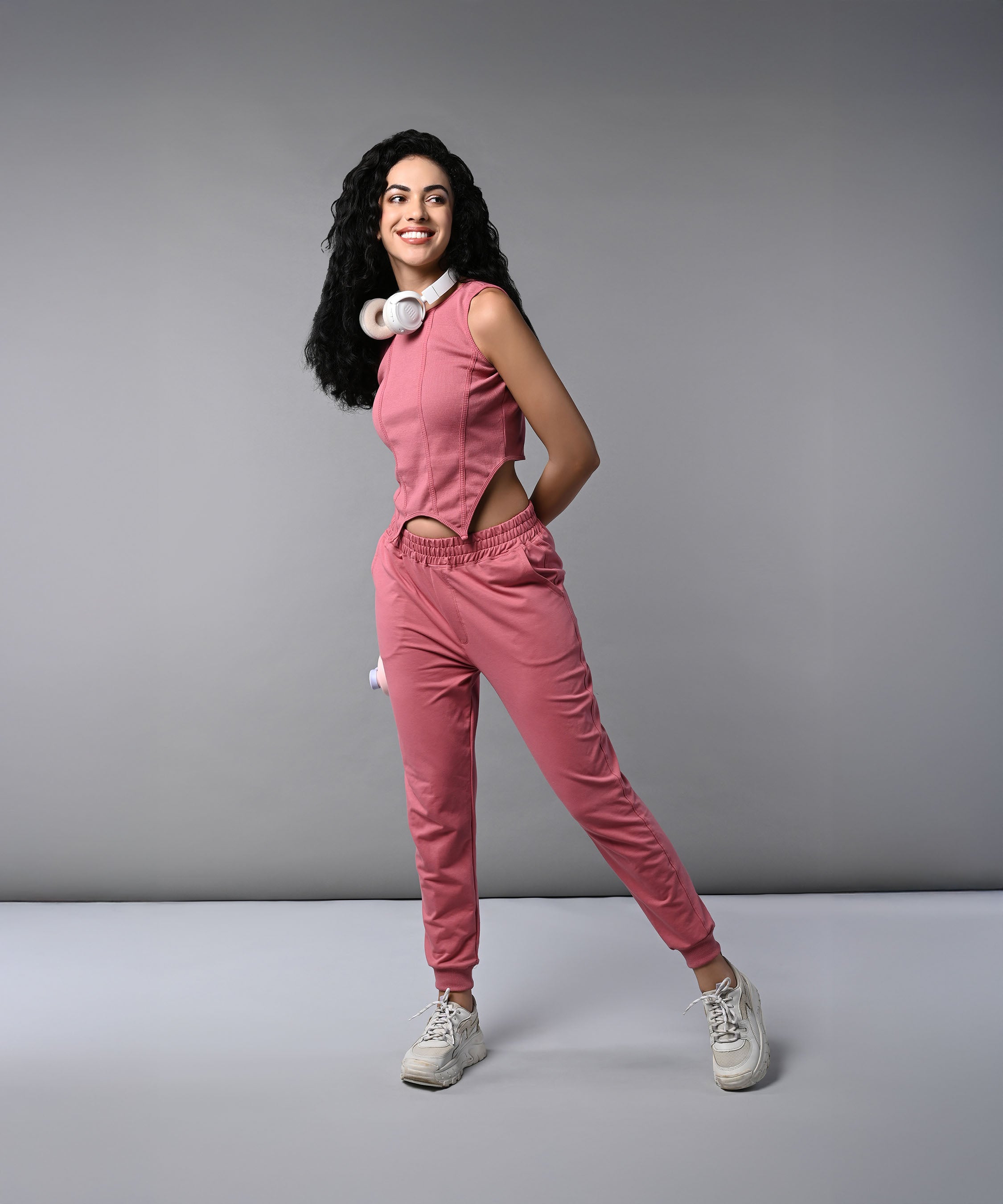 Woman in a pink corset crop-top with matching joggers and headphones around her neck, smiling and posing against a gray background.