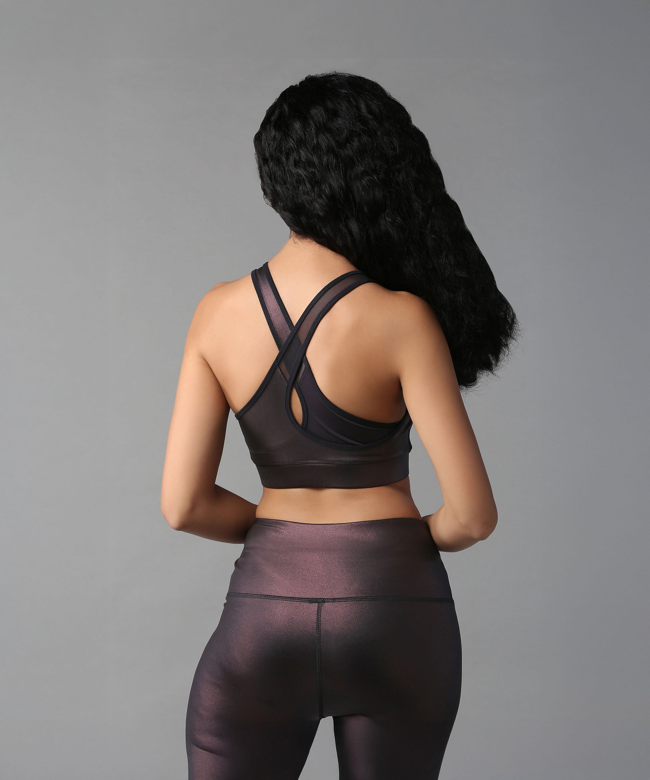 A woman in the Radiant Maroon Shimmer Work out Ensemble with a cross body racerback design is pictured from behind, her long wavy hair visible against a plain background.