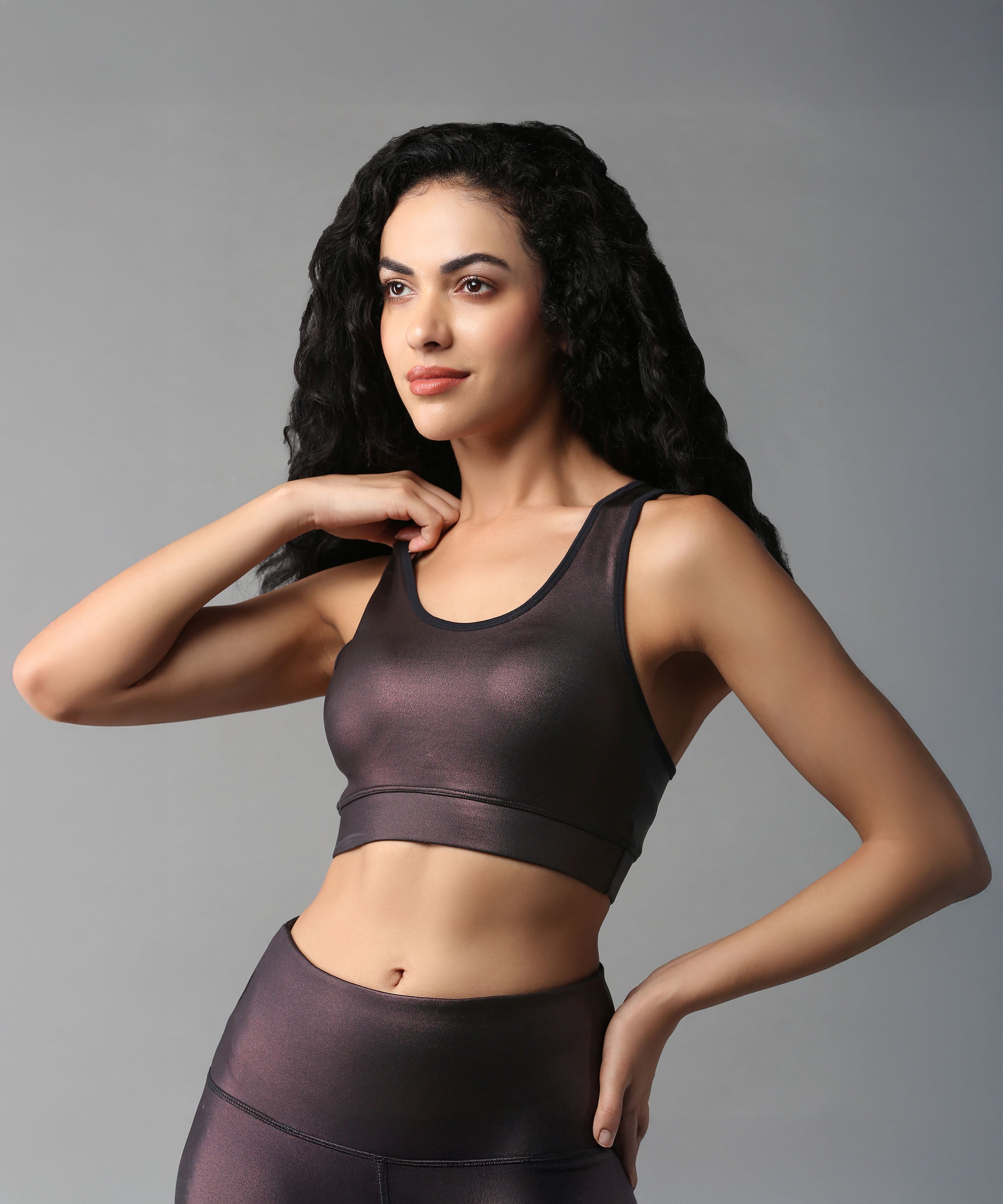 A woman with curly hair poses against a gray background wearing the Radiant Maroon Shimmer Work out Ensemble, featuring a dark crossbody racerback sports bra and moisture-wicking leggings.