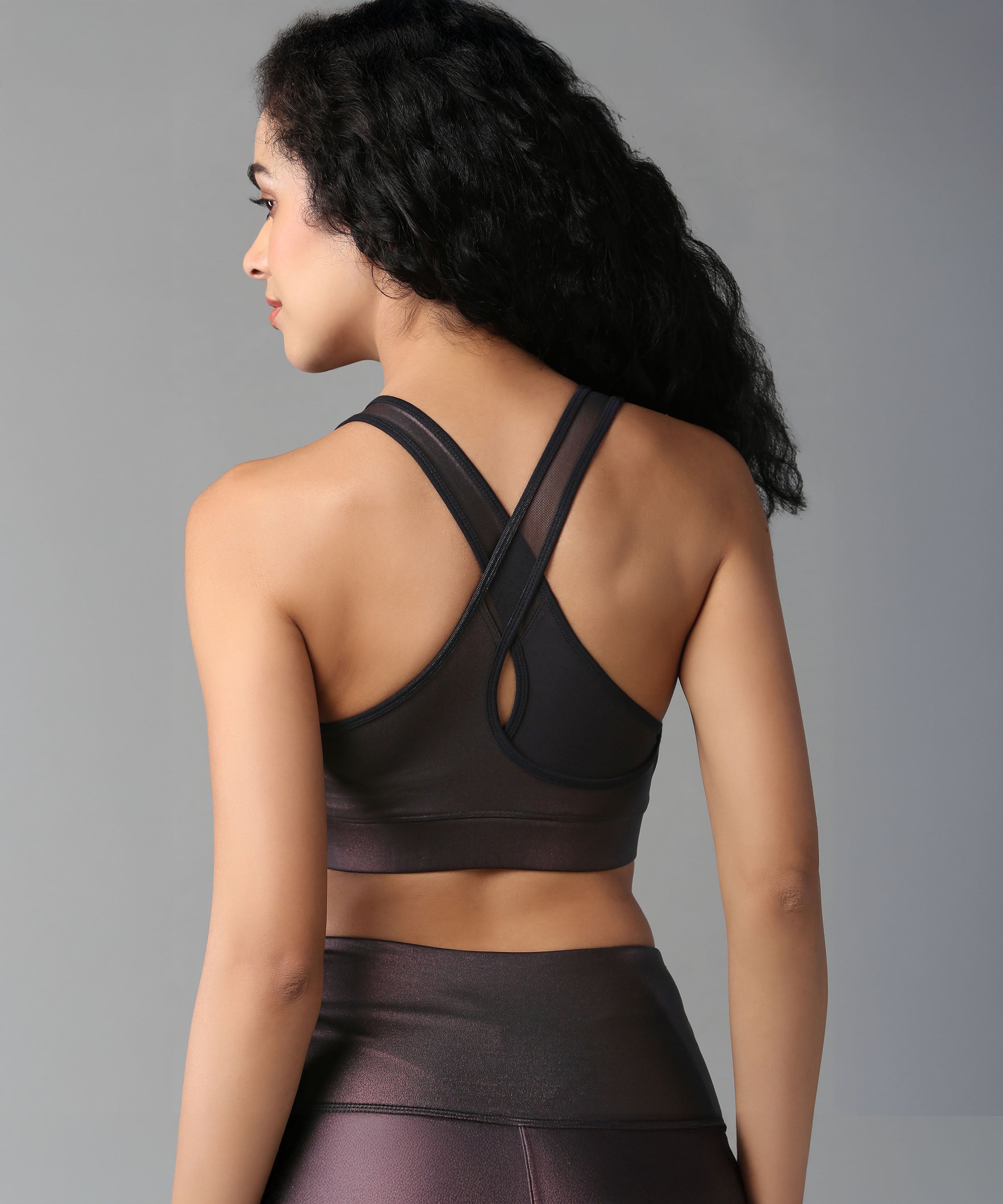 A woman with curly hair models the Radiant Maroon Shimmer Workout Ensemble—sports bra and leggings—in shiny fabric, shown from the back against a gray background.