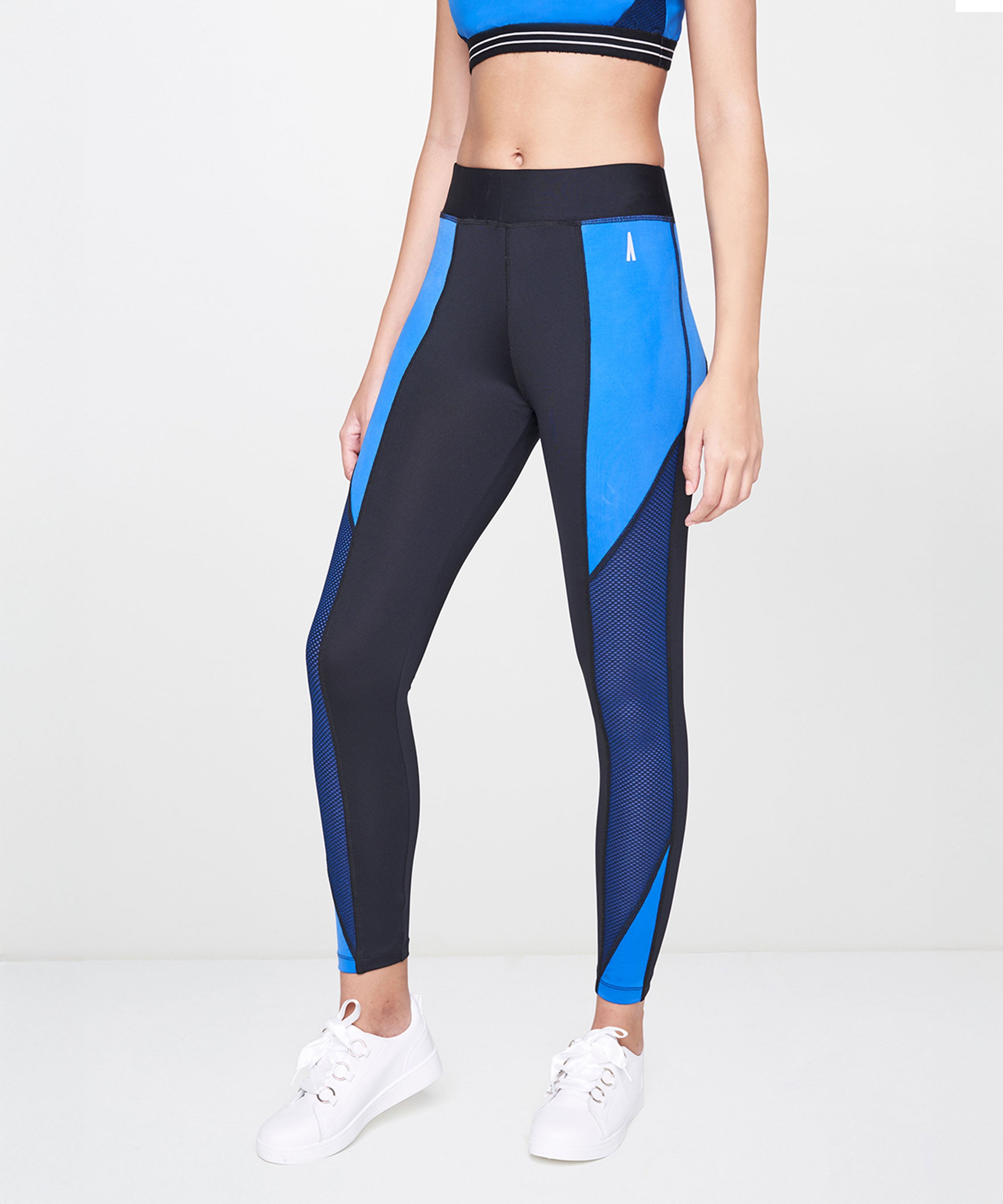 A woman models Indigo Leggings and a matching sports bra with white sneakers against a plain background.