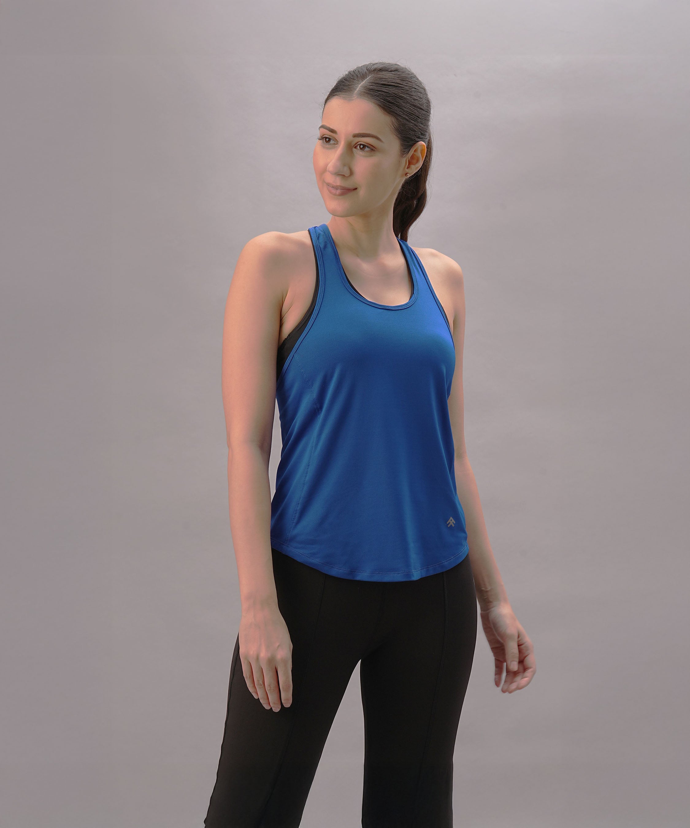 A woman models the Blue Racer Back Top and black leggings against a plain gray background, highlighting stylish activewear.