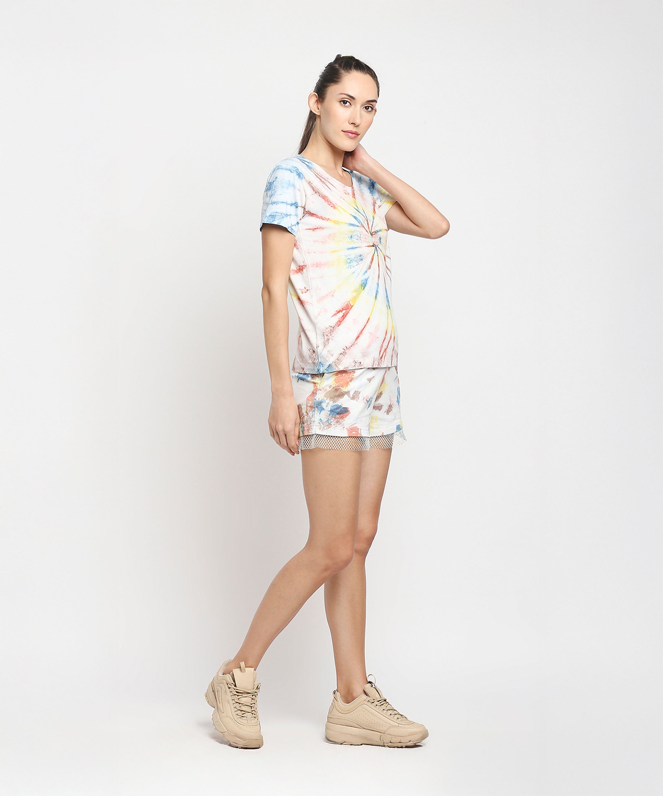 Woman poses against a plain white background wearing the Netted shorts + comfort tee in colorful tie-dye, paired with beige sneakers. Eco-friendly dyes give her cotton outfit vibrant flair.