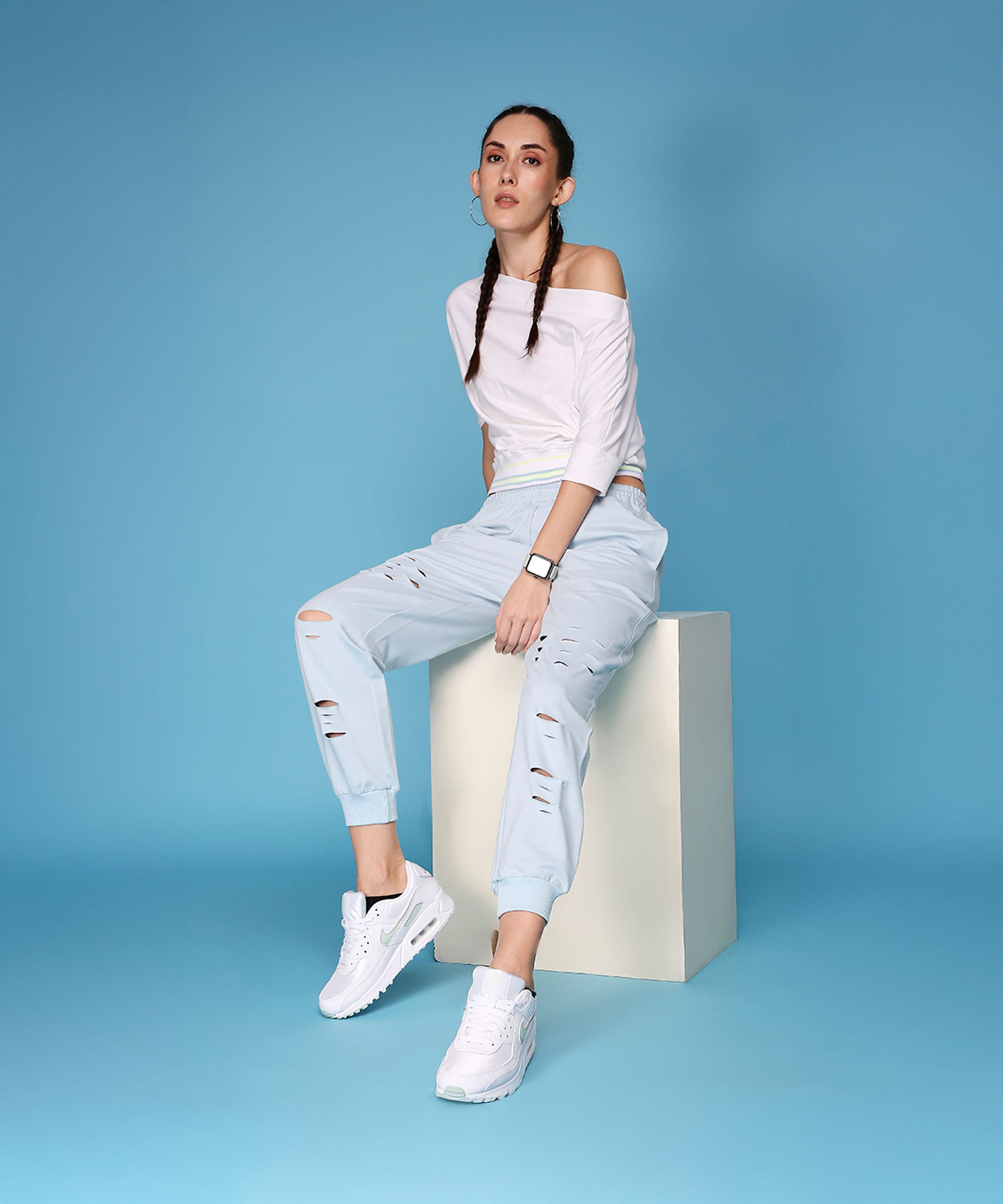 A woman models the AW21 Off Shoulder White Rib top and Light Blue Ripped Joggers while sitting on a cube against a blue background.