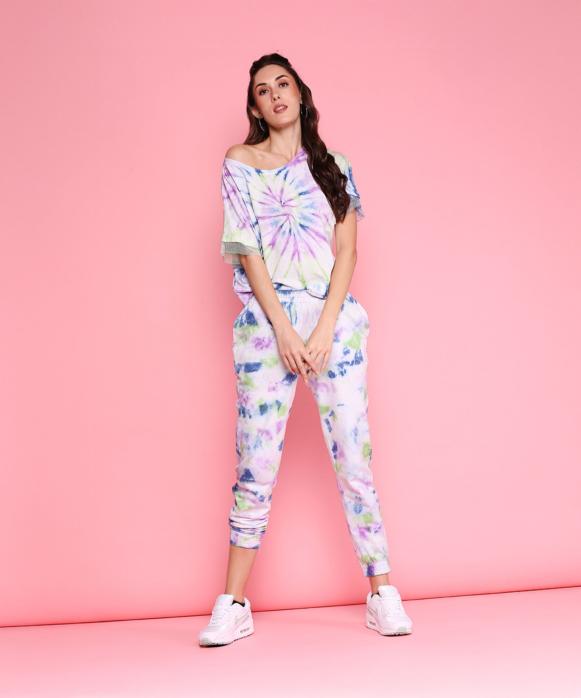 A woman wearing the Comfort Fit Tee + Joggers - AW21 lounge set and white sneakers poses against a pink background.