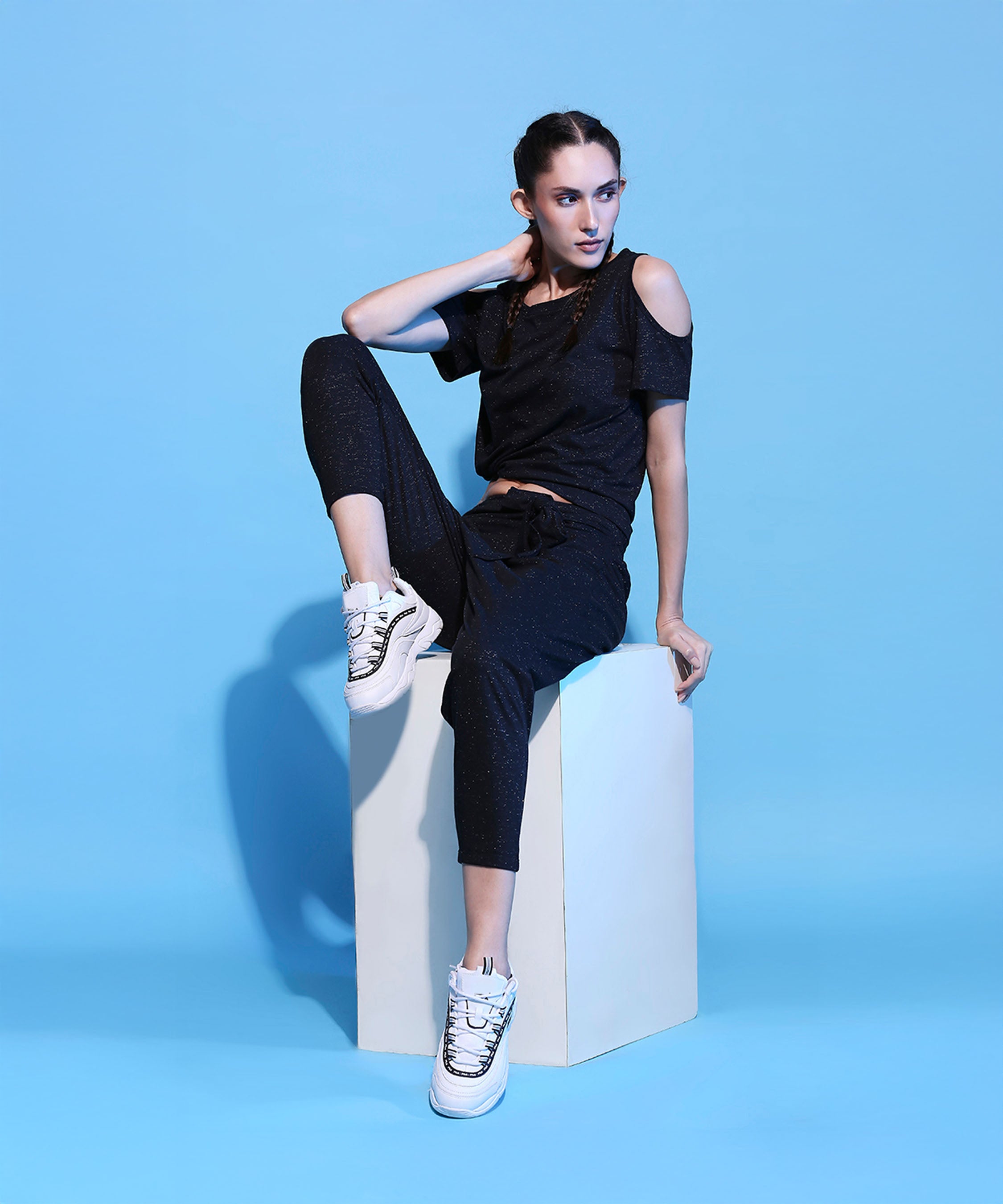 A woman in the Shimmer Cold Shoulder Tee (Customised) and Side Tie Up Shimmer Yoga Pants- AW21 with white sneakers sits on a cube against a blue background, highlighting the sleek, cotton-blended shimmer fabric.